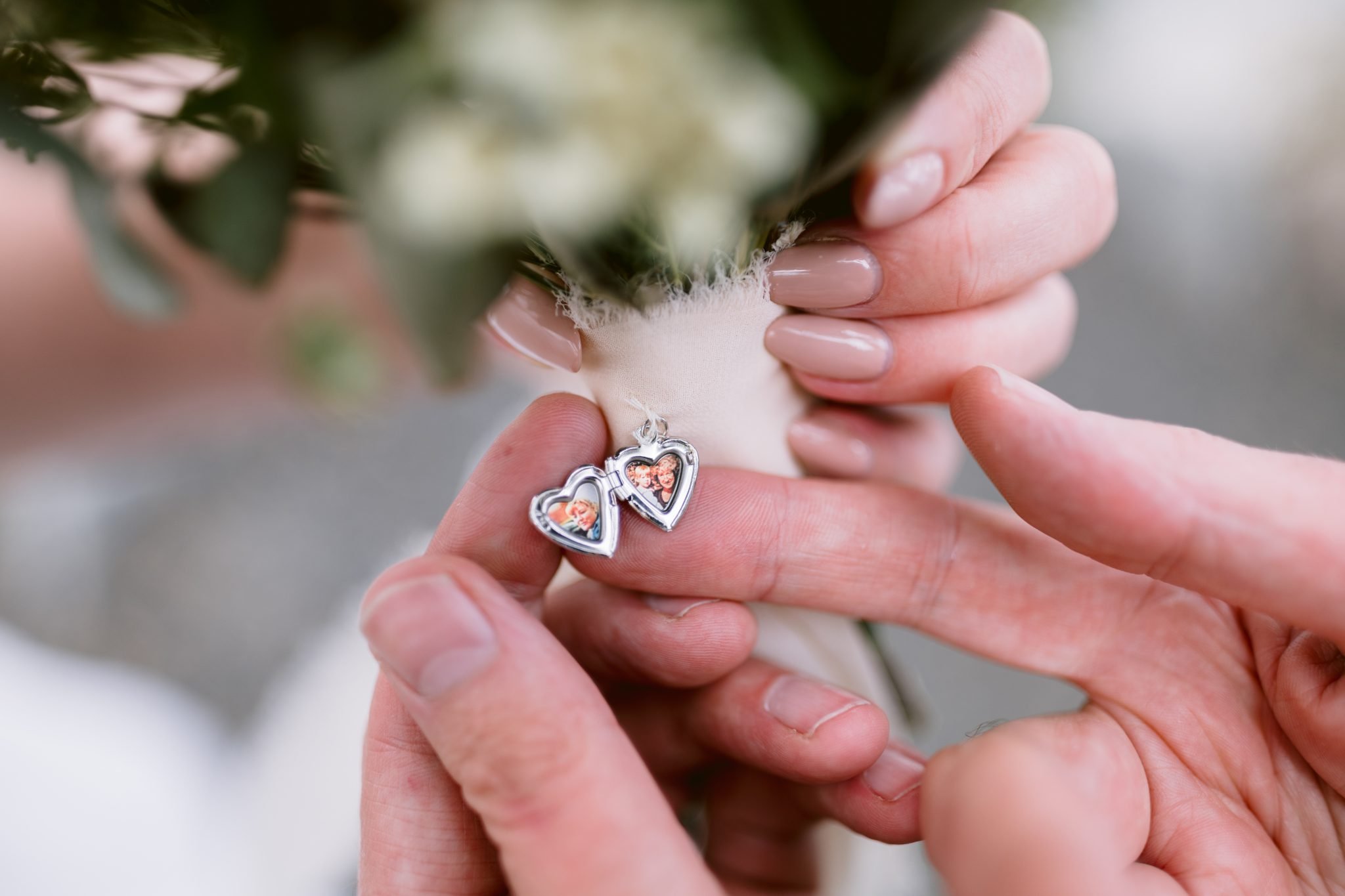 Hands holding a heart-shaped locket featuring small photos, attached to a bouquet wrapped in ribbon.