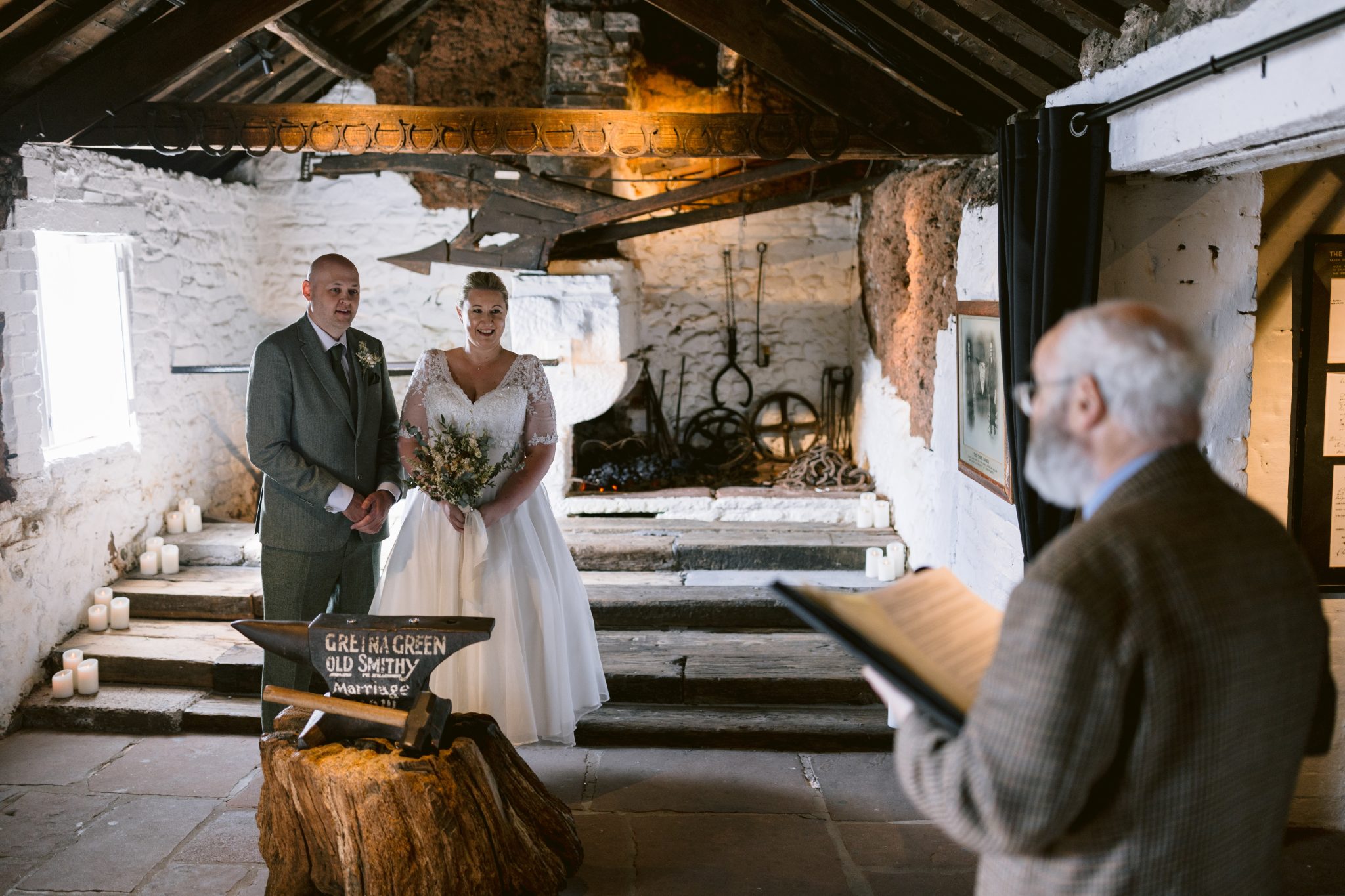 A couple stands in a rustic venue during a wedding ceremony, with an officiant speaking. Candles are lit around the room.