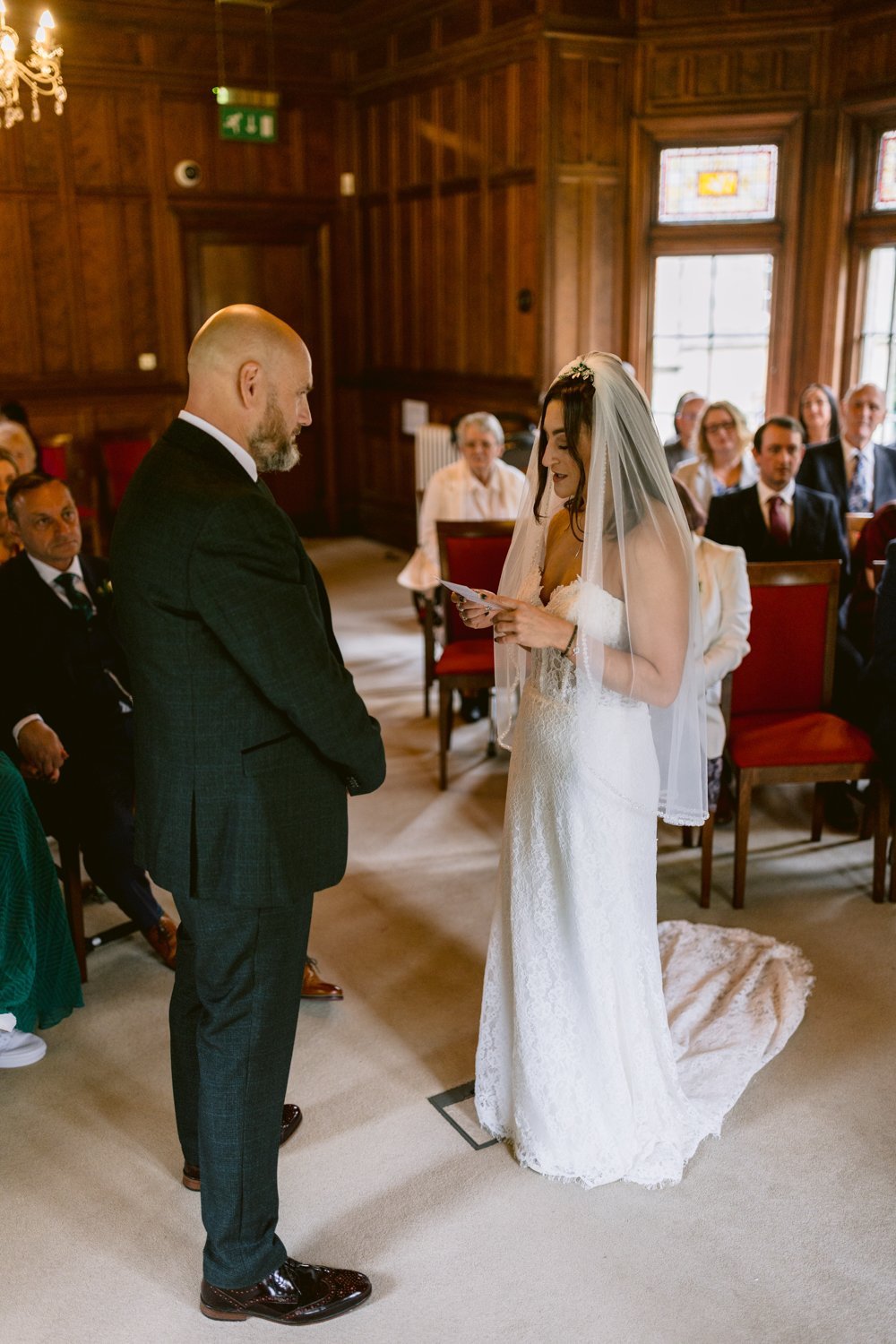 In the elegant setting of Lady Gilford’s House, the bride and groom stand facing each other in a wood-paneled room, exchanging vows as seated guests watch.