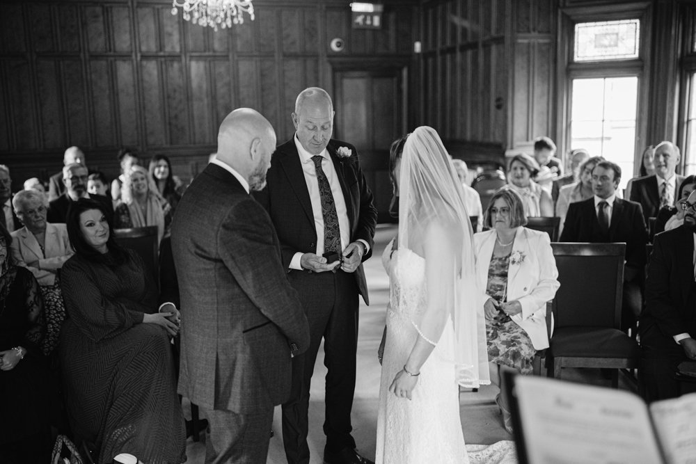 In a wood-paneled room at Lady Gilford's House, a bride and groom stand with an officiant, facing their seated guests during the wedding ceremony.