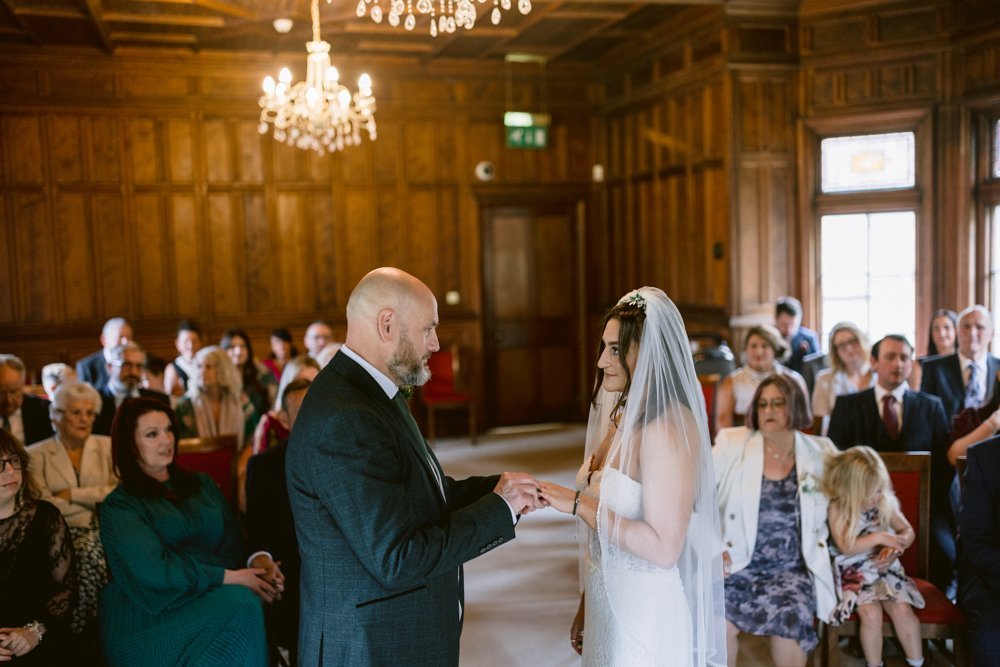In the elegant wood-paneled room of Lady Gilford’s House, a couple exchanges rings under sparkling chandeliers, while guests seated in rows witness the beautiful ceremony.