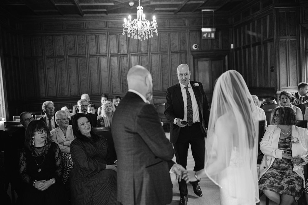A bride and groom stand in a wood-paneled room at Lady Gilford’s House, holding hands as they face the officiant. Guests are seated, watching the ceremony unfold beneath a sparkling chandelier. The timeless scene is captured in black and white.