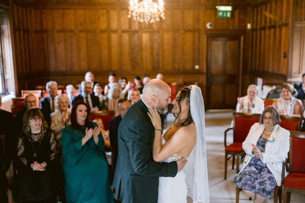 In the elegant setting of Lady Gilford’s House, a couple embraces at their wedding ceremony in a wood-paneled room, with seated guests smiling and applauding.