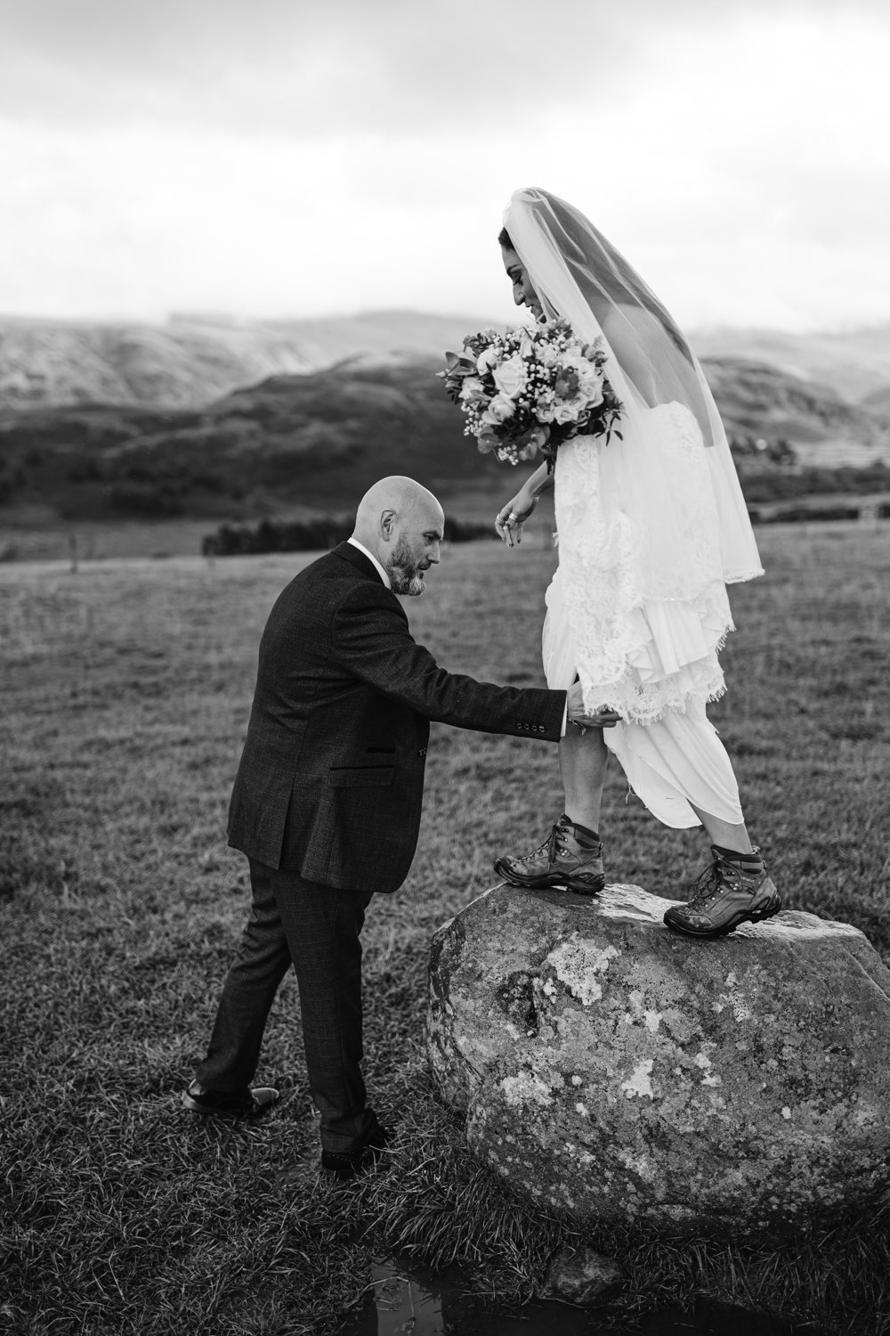 Amidst the rolling hills, a bride stands confidently on a rock, her hiking boots peeking beneath her gown. Nearby, Lady Gilford’s House adds an air of timeless elegance as the groom gently steadies her leg in support.