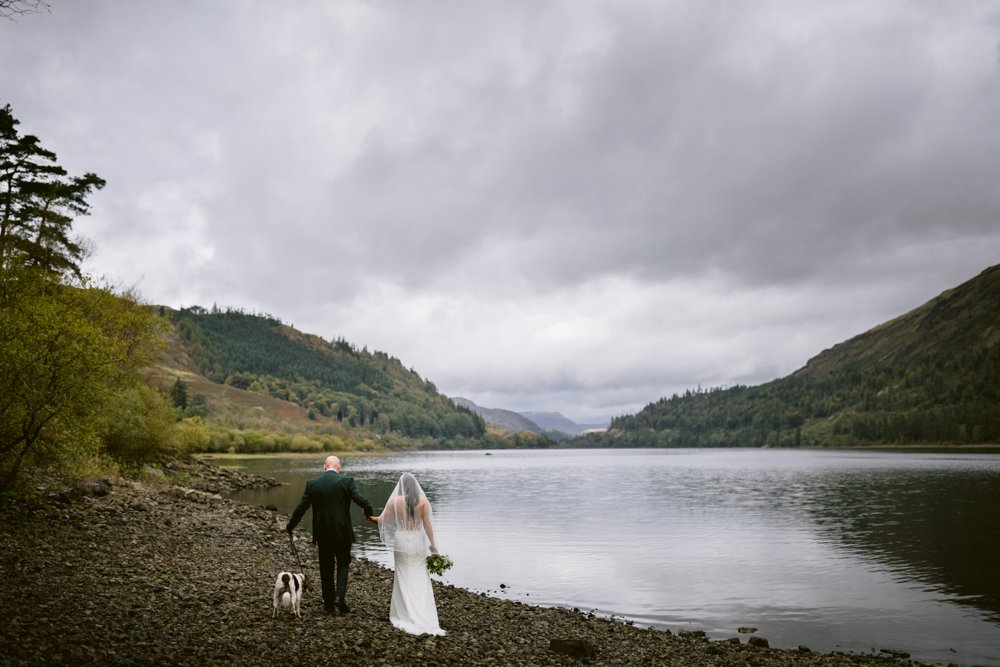 A bride and groom walk hand in hand with a dog along a rocky lakeside near Lady Gilford’s House, under a cloudy sky, surrounded by hills and trees.