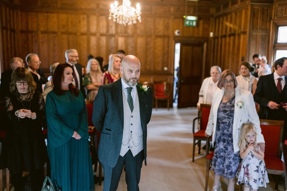 Amidst the elegant ambiance of Lady Gilford’s House, a man in formal attire stands at the front of a wood-paneled room. Around him are seated and standing guests, including a woman with a young girl. A chandelier gracefully illuminates the gathering.