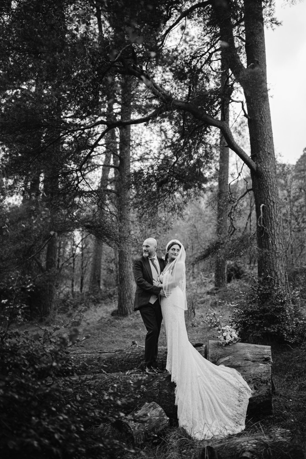 A bride and groom stand on a fallen log in the forest near Lady Gilford’s House, both smiling. The bride, in a long white dress and veil, holds a bouquet while the groom dons a suit. Tall trees envelop them in this enchanting setting.