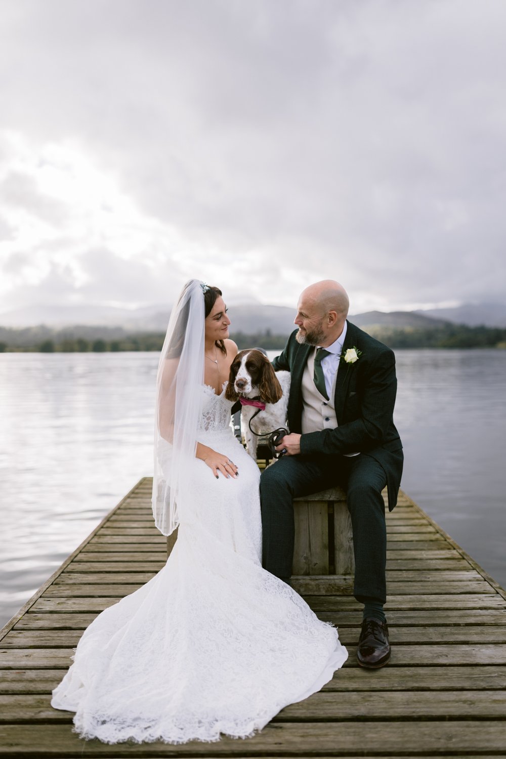 Bride and groom sit on a dock with their dog by the lake at Lady Gilford’s House. The bride is clad in her white gown and veil, while the groom dons a suit. An overcast sky and calm water provide a serene backdrop to this picturesque moment.