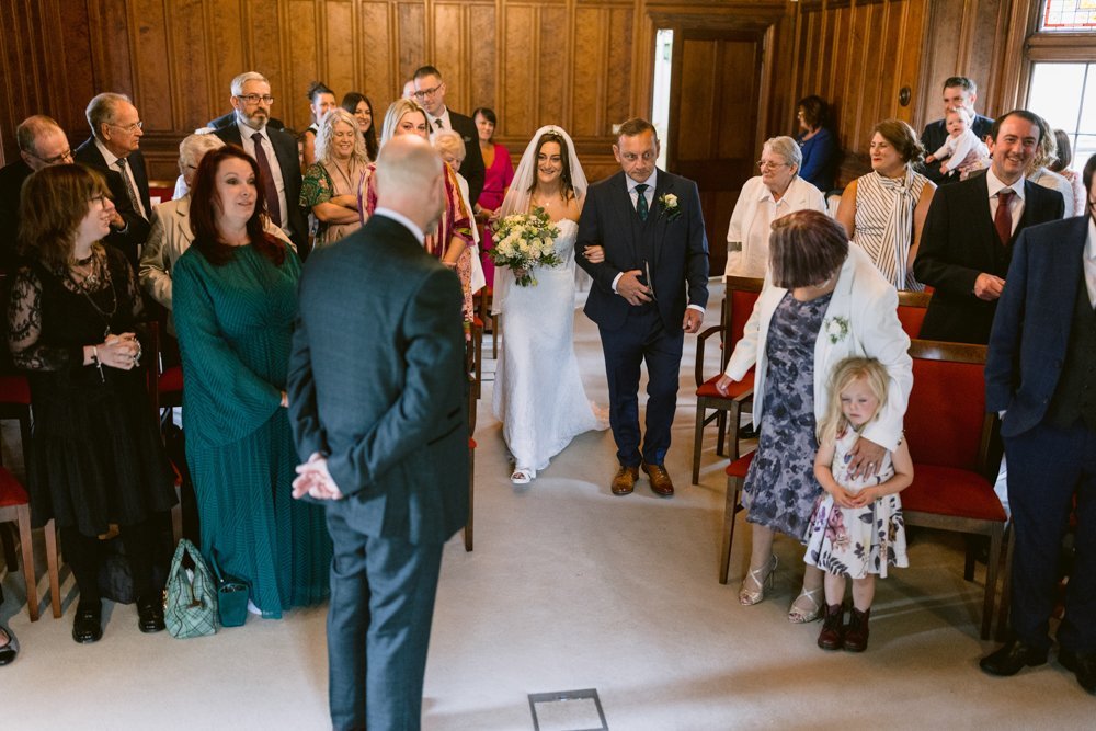 In the elegant wood-paneled room of Lady Gilford’s House, the bride gracefully walks down the aisle with a man by her side, as guests watch in awe, some seated and others standing, capturing this beautiful wedding ceremony.