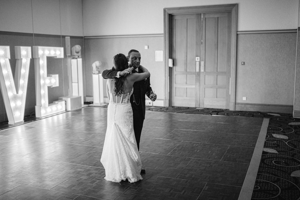 A bride and groom share a dance on the wooden floor of Lady Gilford’s House, surrounded by elegant decor and a large lit "LOVE" sign glowing warmly in the background.