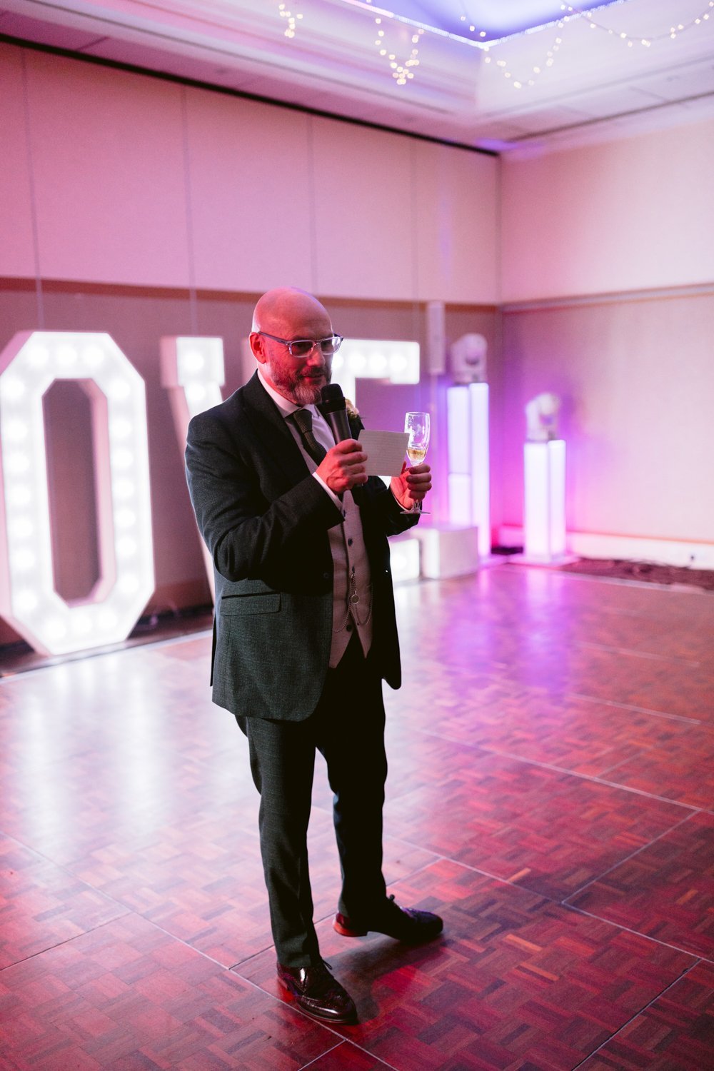 A man in a suit stands on the dance floor of Lady Gilford's House, holding a microphone and a glass, illuminated letters casting a glow in the background.