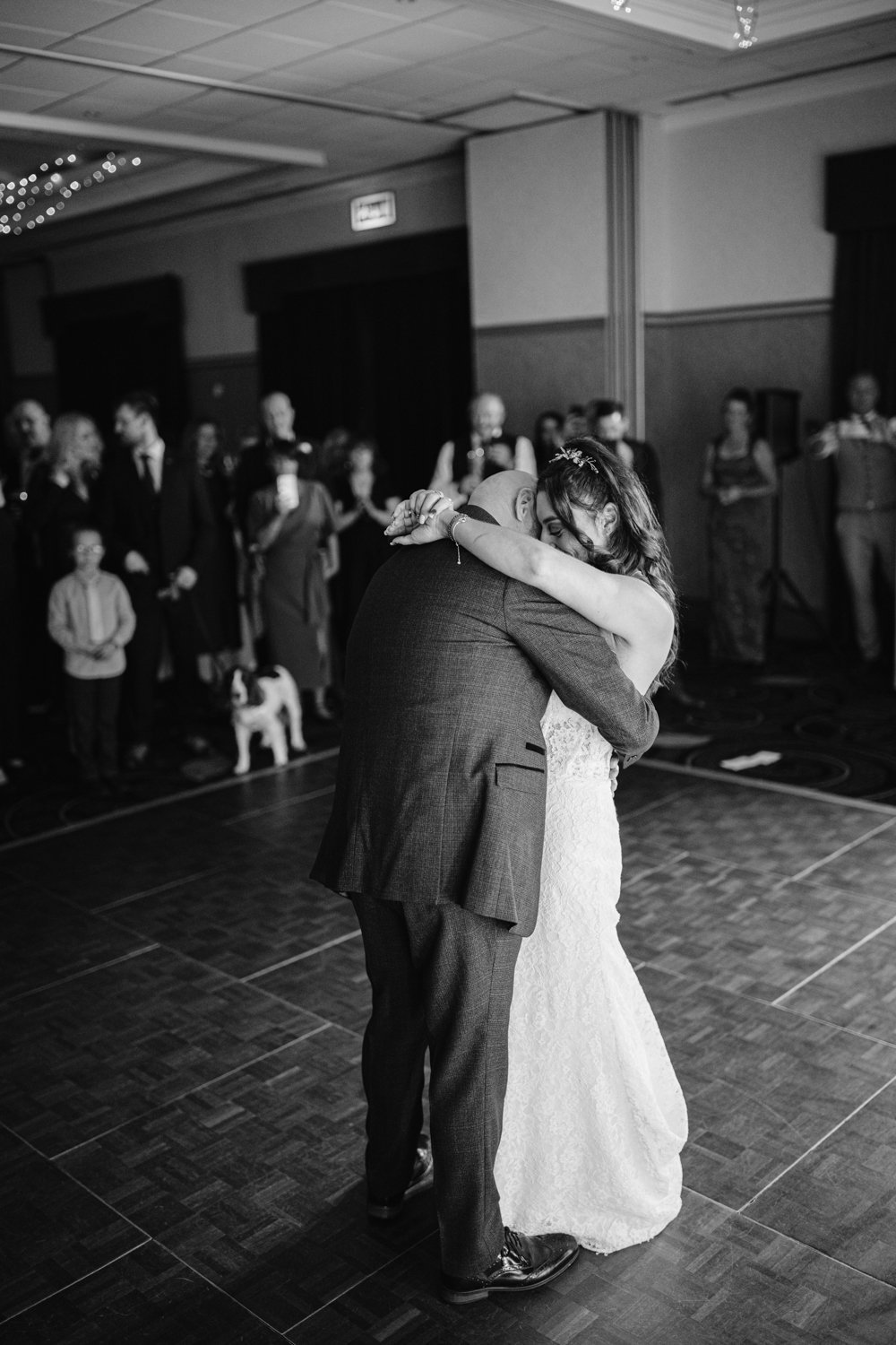 In the grand ballroom of Lady Gilford’s House, a bride and groom share a slow dance. Guests surround them, capturing the moment with photos, while a small dog stands nearby, adding charm to the elegant scene.
