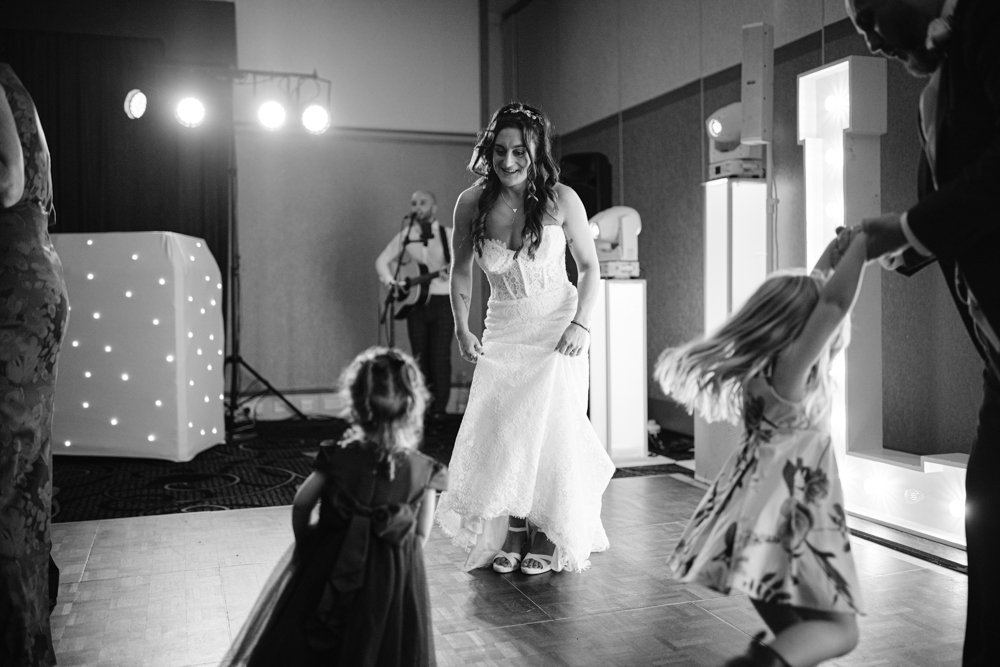 At Lady Gilford’s House, the bride shares a joyful dance with two young girls in dresses, illuminated by vibrant lights with a musician playing sweet melodies in the background.