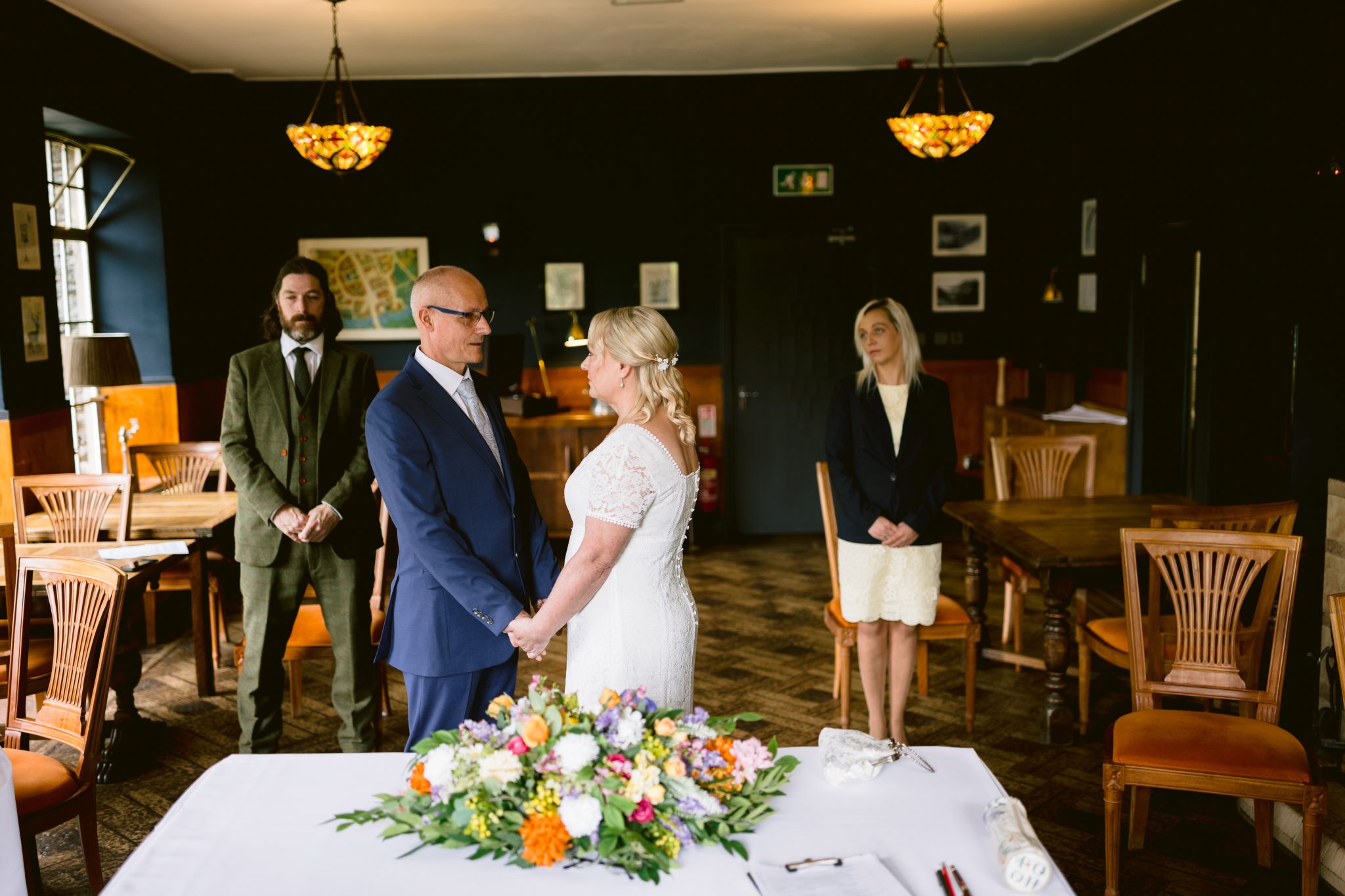 A couple stands facing each other, holding hands in a decorated room, with two people standing in the background.