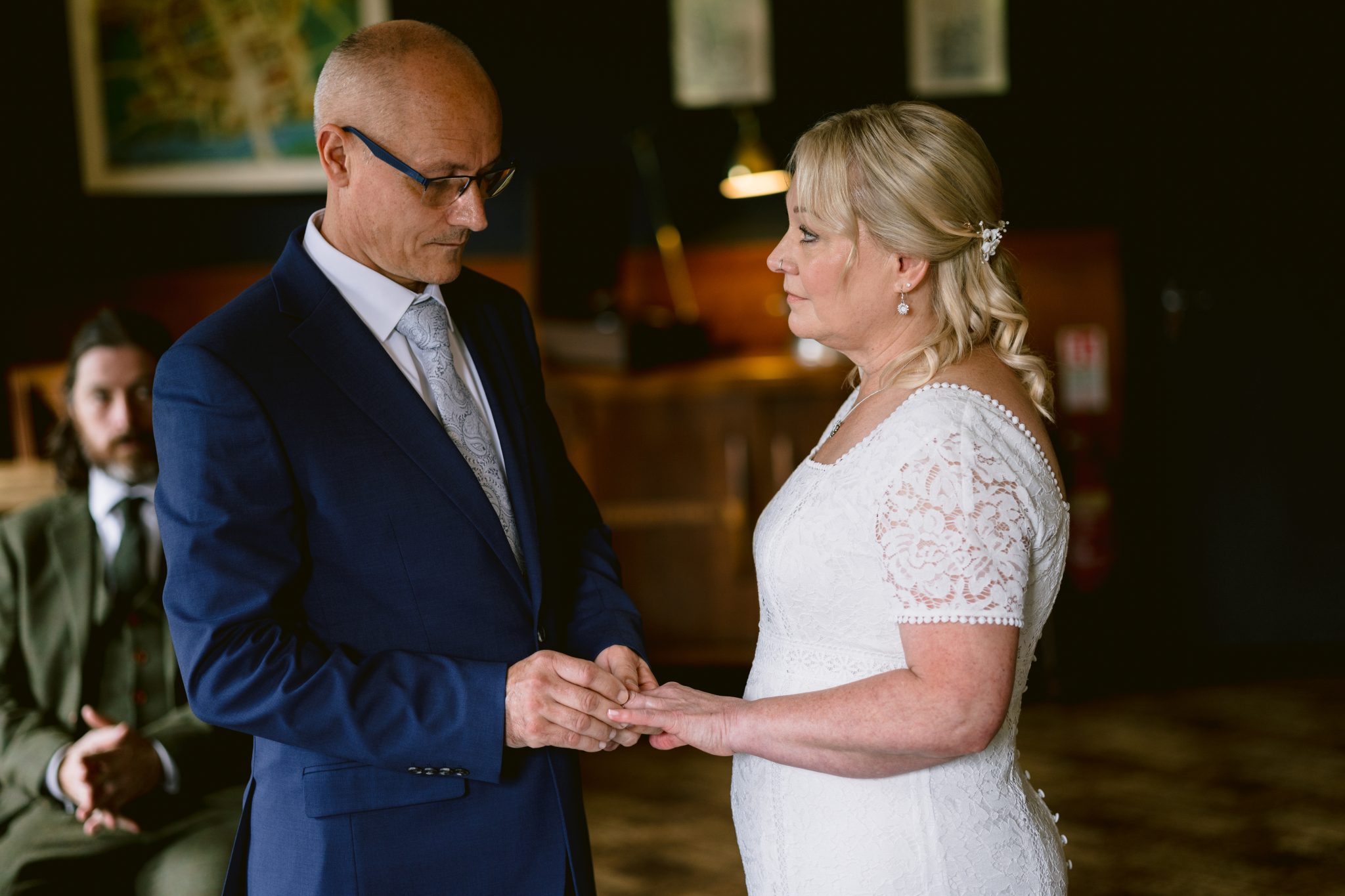 A couple exchanges rings during a wedding ceremony, with a seated man in the background.