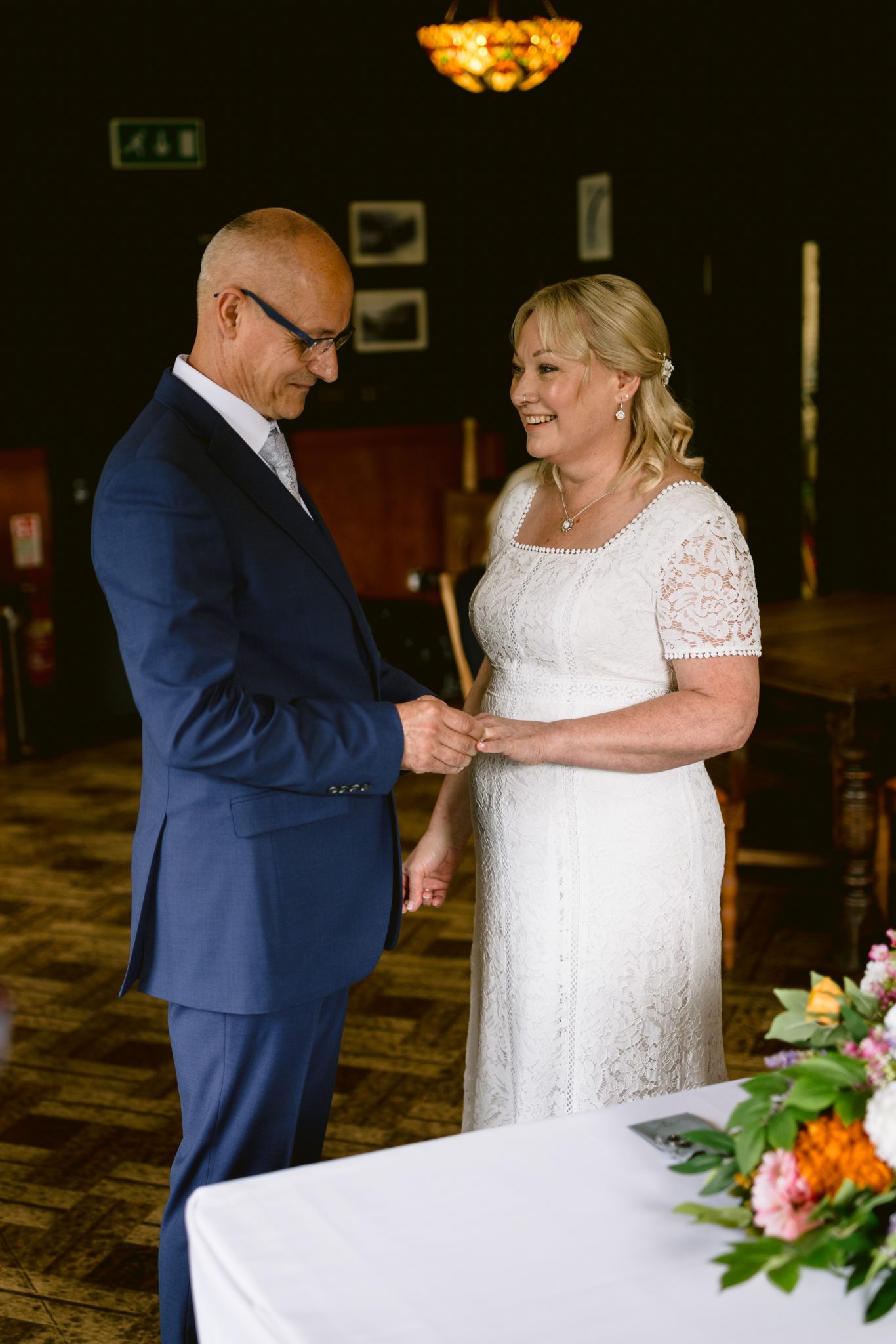 A couple stands together in formal attire, holding hands and smiling, in what appears to be a wedding setting with a bouquet in the foreground.