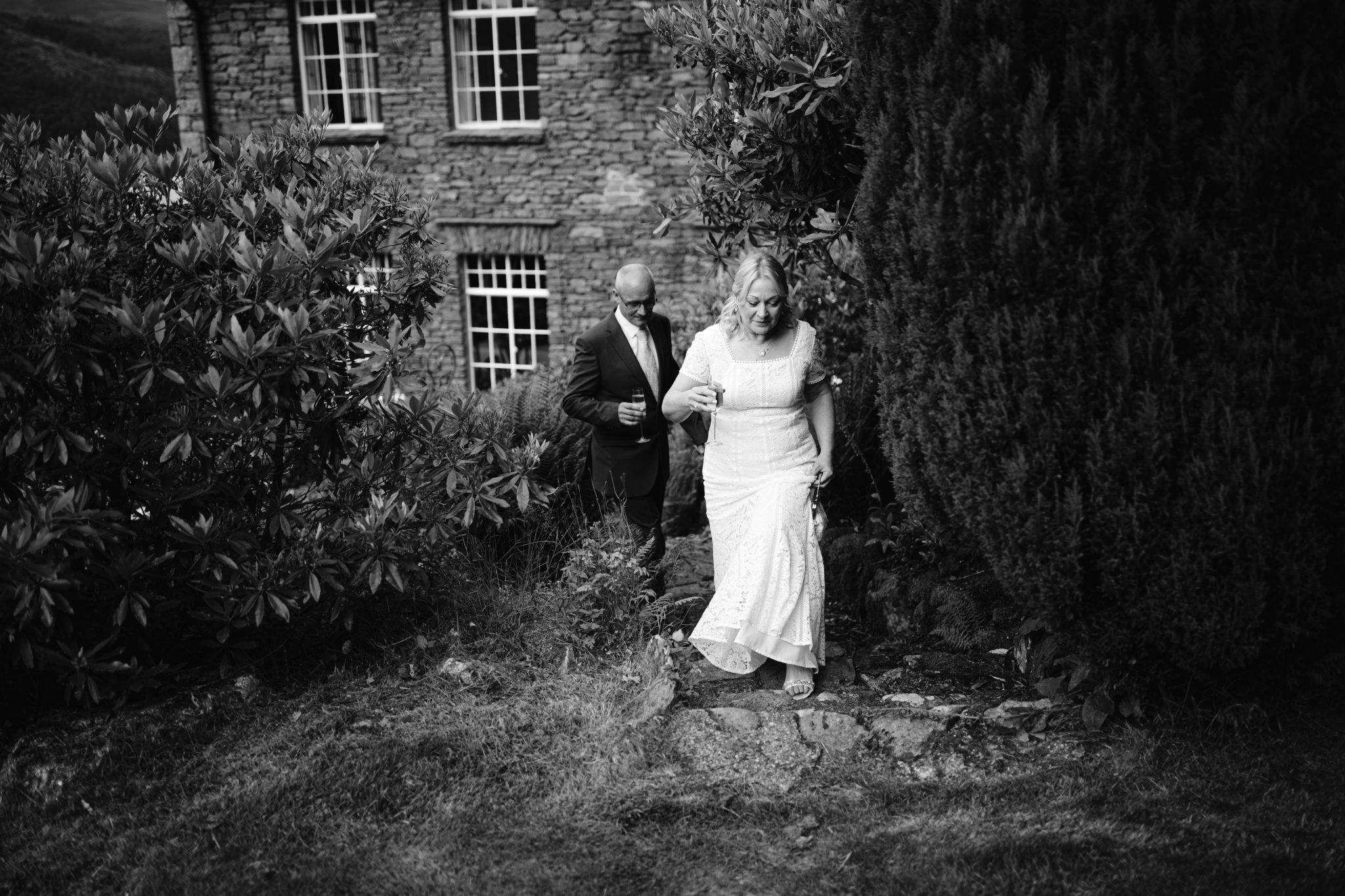 A bride in a white dress walks down garden steps, followed by a man in a dark suit. A brick house with large windows is in the background.