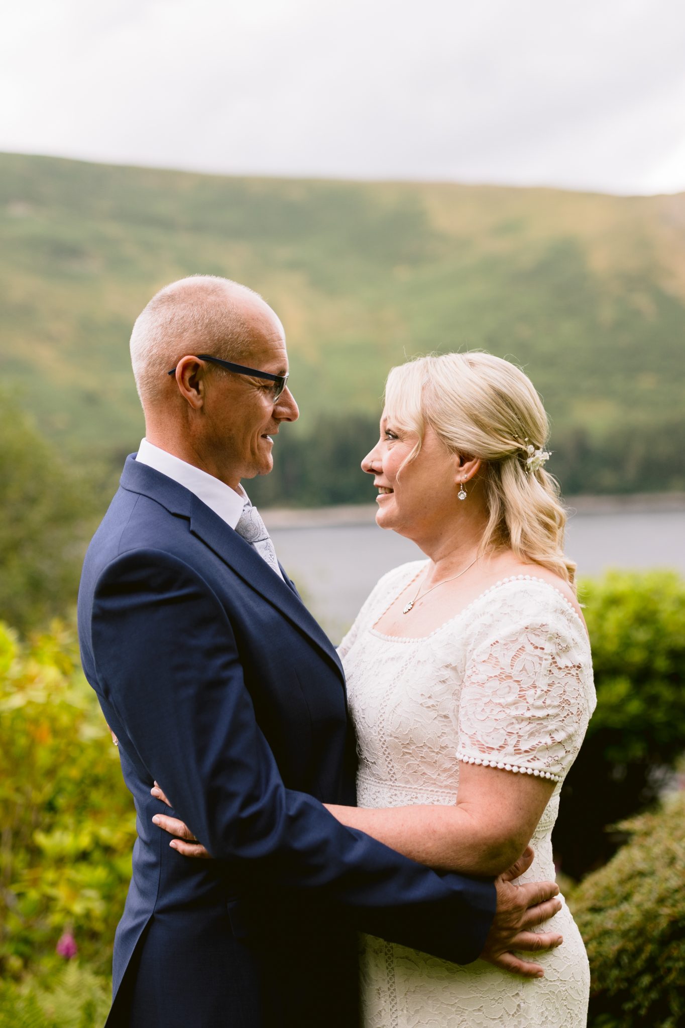 A couple dressed in formal attire embrace and smile at each other outdoors, with a scenic view of hills and water in the background.