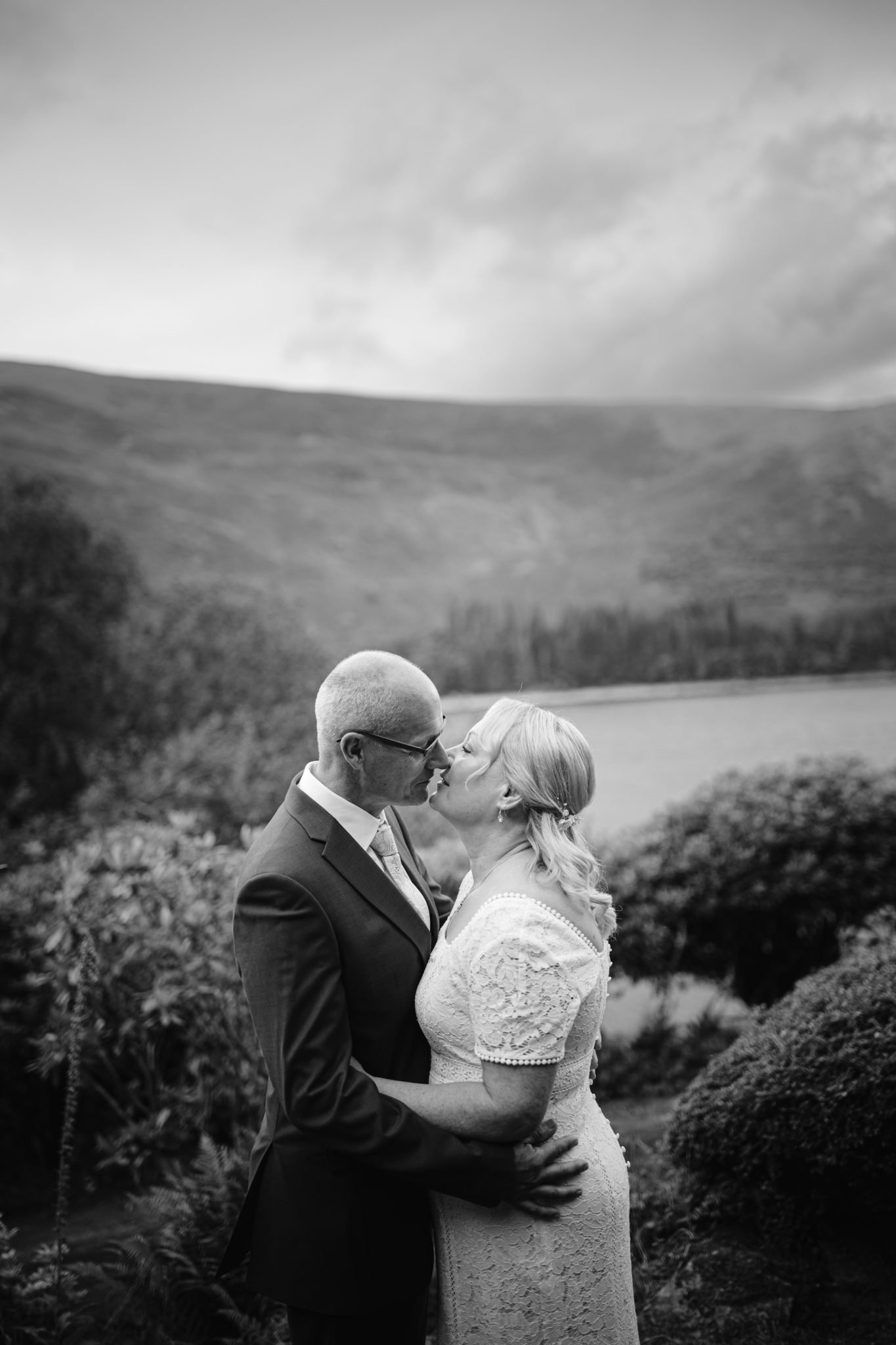 A couple in formal attire kisses outdoors with a lake and mountains in the background, in black and white.