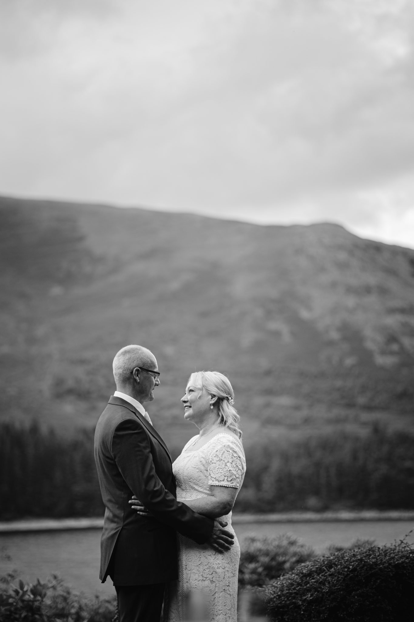 A couple in formal attire stands embracing and gazing at each other, with a mountainous landscape in the background. Black and white image.