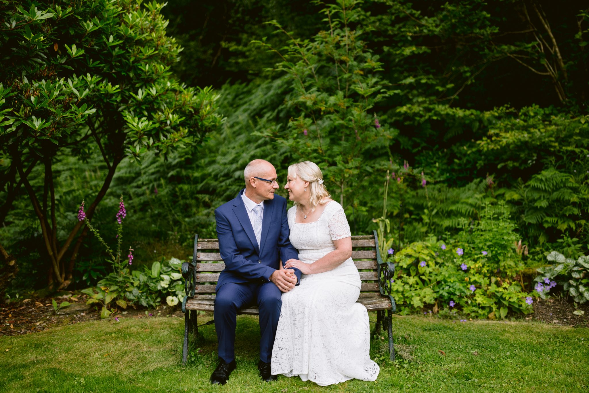 An older couple sits on a wooden bench in a garden. The man is in a blue suit, and the woman wears a white dress. They hold hands and face each other, surrounded by greenery and flowers.