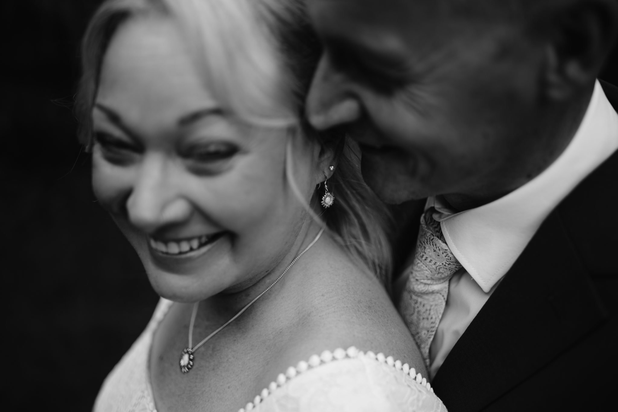 A black-and-white close-up of a smiling couple in formal attire. The woman is wearing earrings and a necklace.