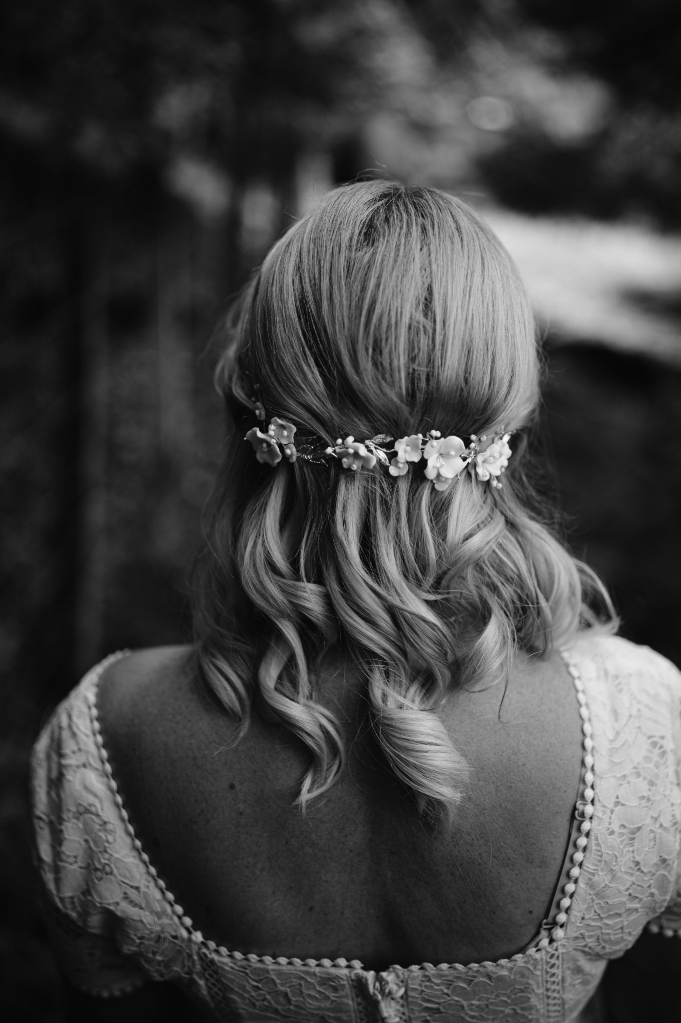 A woman with wavy hair faces away, wearing a floral hairpiece and a lace dress. The image is in black and white.