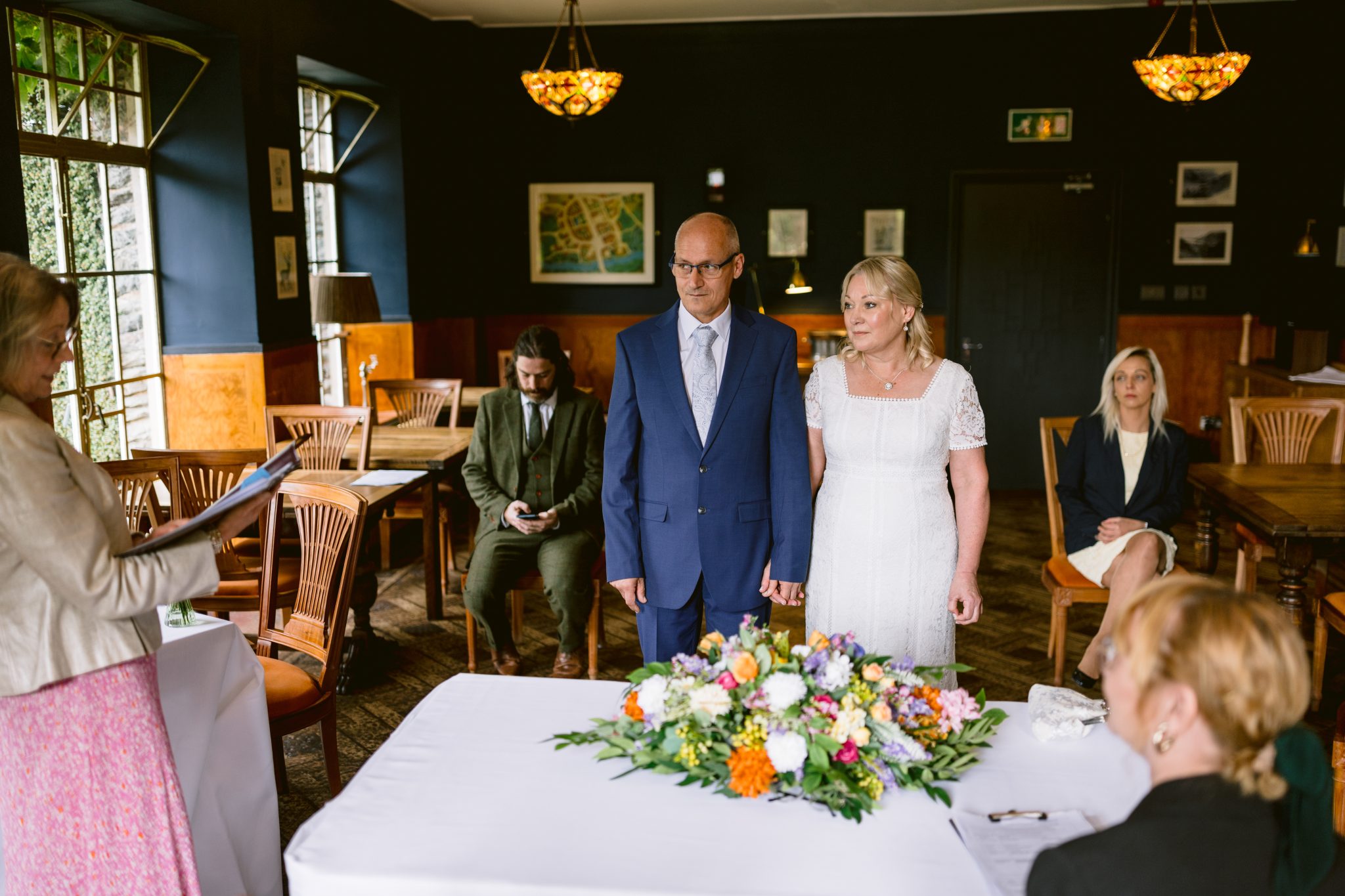 A couple stands at the front of a room during a ceremony, with a floral arrangement on the table. Three people are seated in the background.