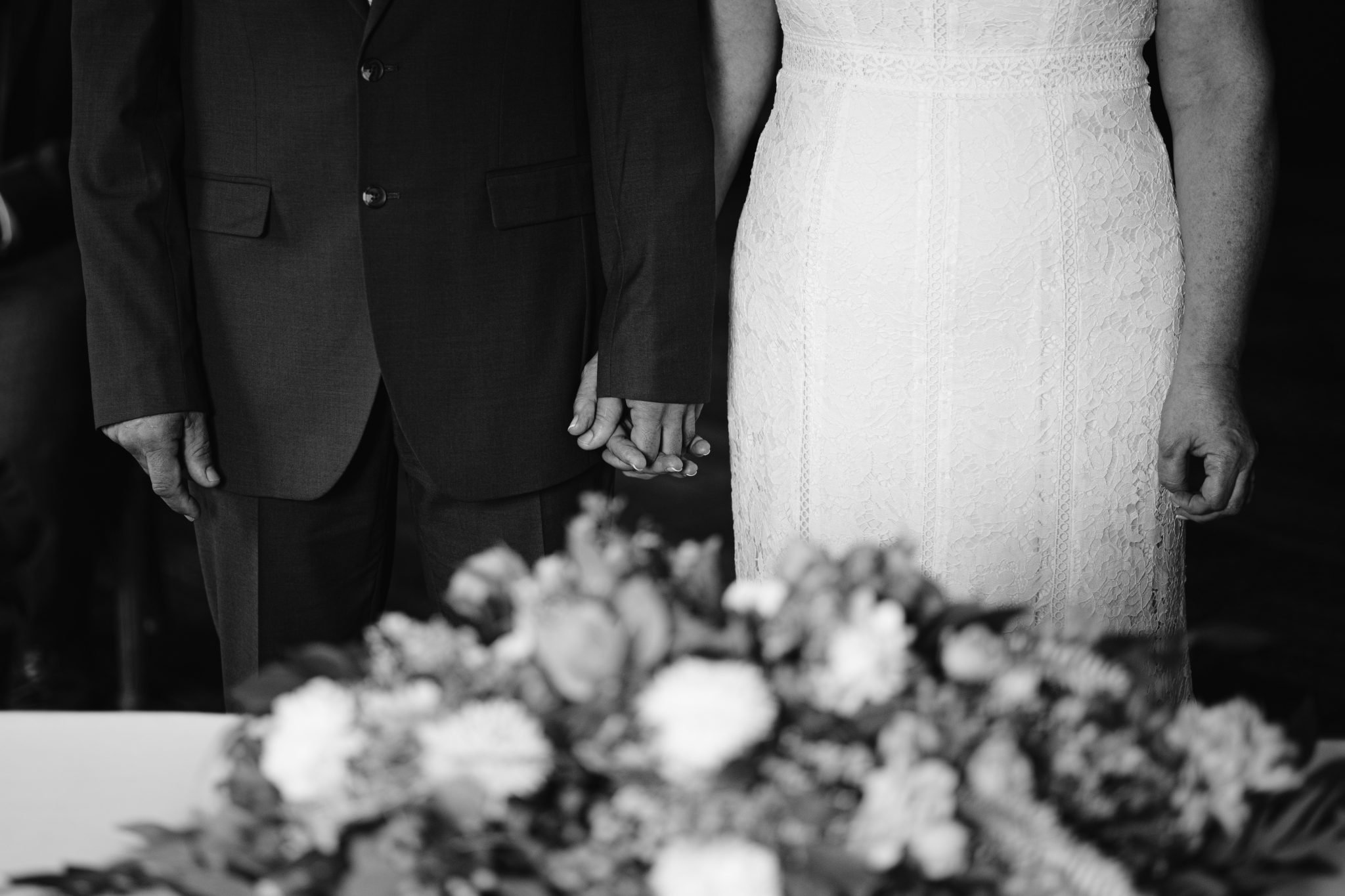 A couple holding hands during a wedding ceremony, with a floral arrangement in the foreground. Black and white image.