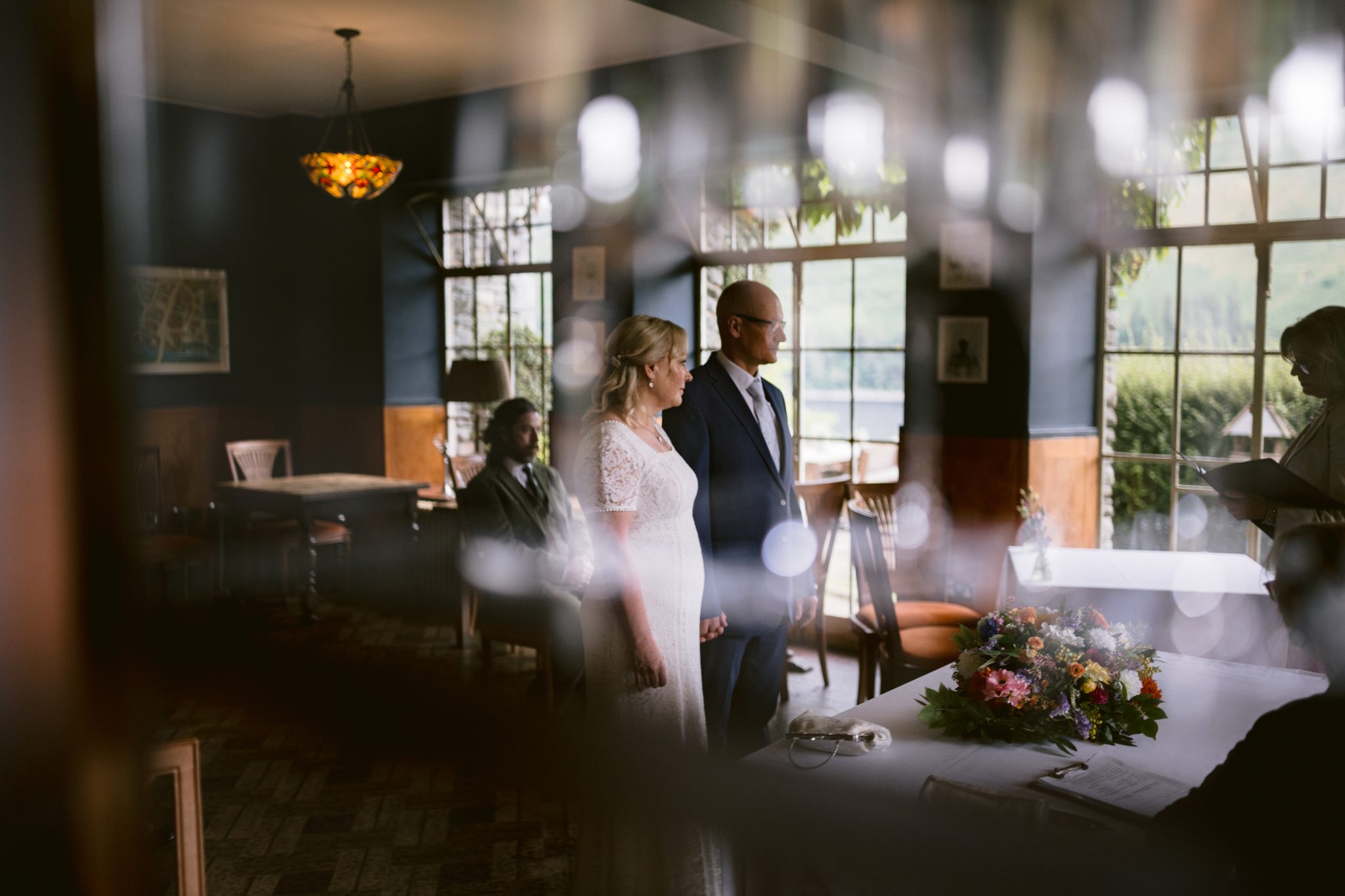 A couple stands in a dimly lit room, apparently during a wedding ceremony, with a bouquet of flowers on a table and a person seated nearby.