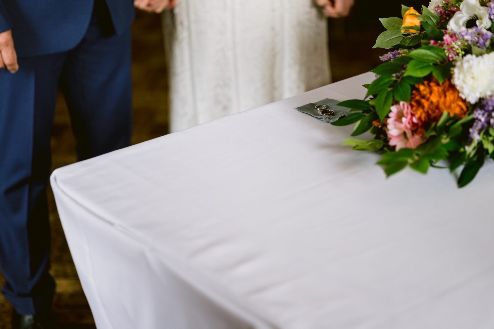 A couple stands by a table with a bouquet and wedding rings on a white cloth.