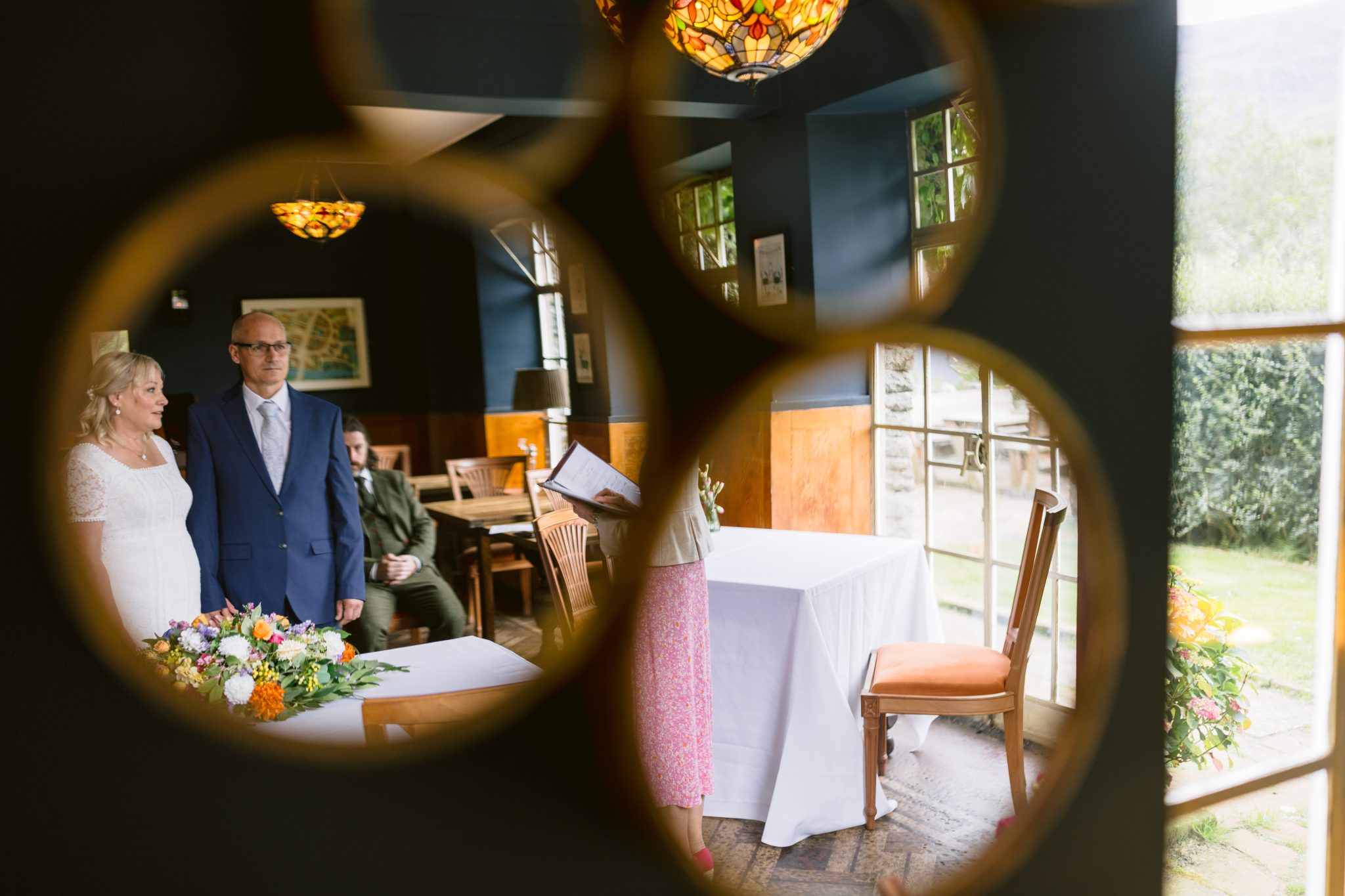 A couple stands inside a room with decorative mirrors, while a person reads from a sheet. A table with flowers and a view of the outdoors can be seen.