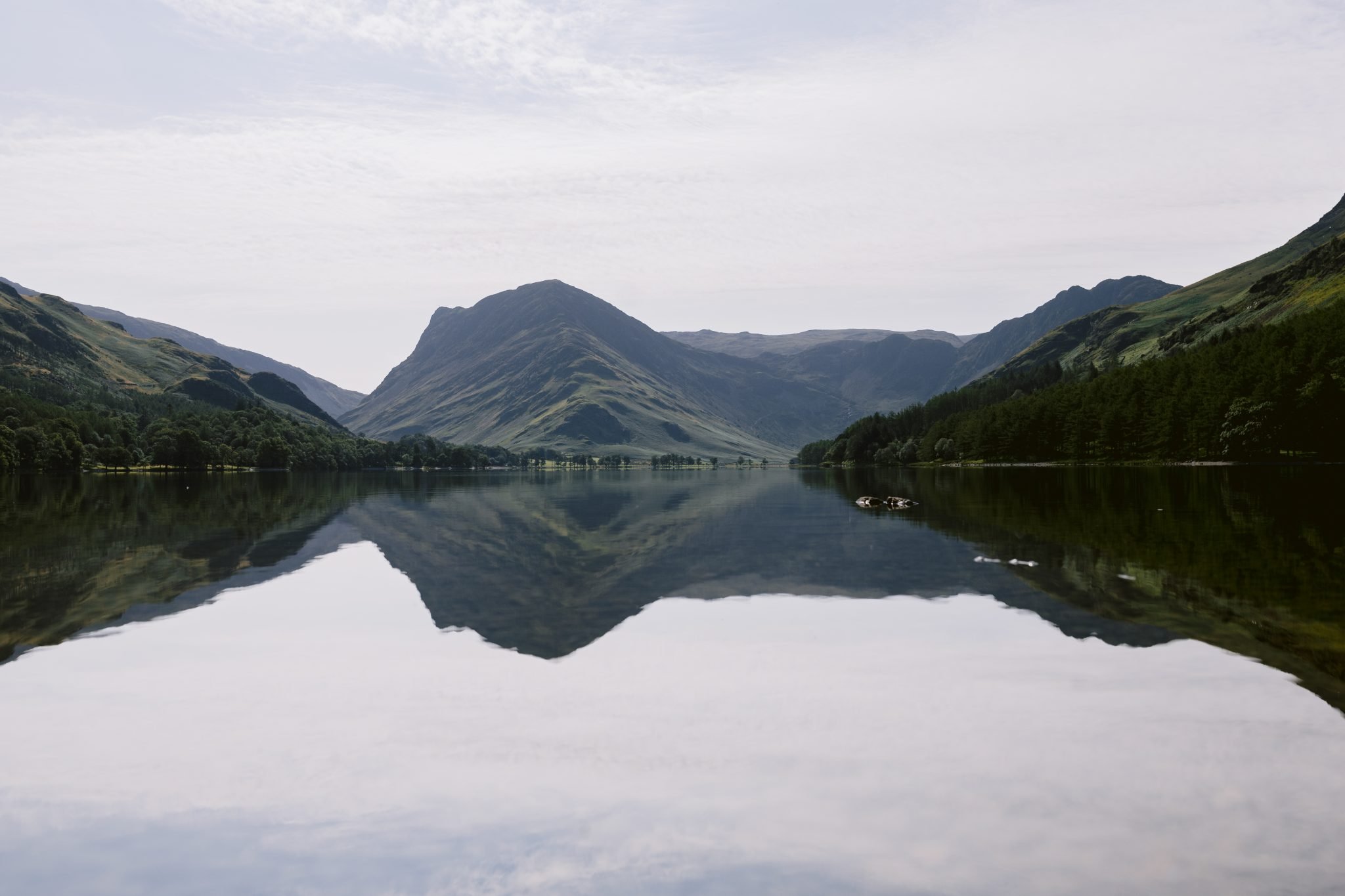 A calm lake reflects surrounding mountains and a cloudy sky, with a tree-lined shore in the distance.