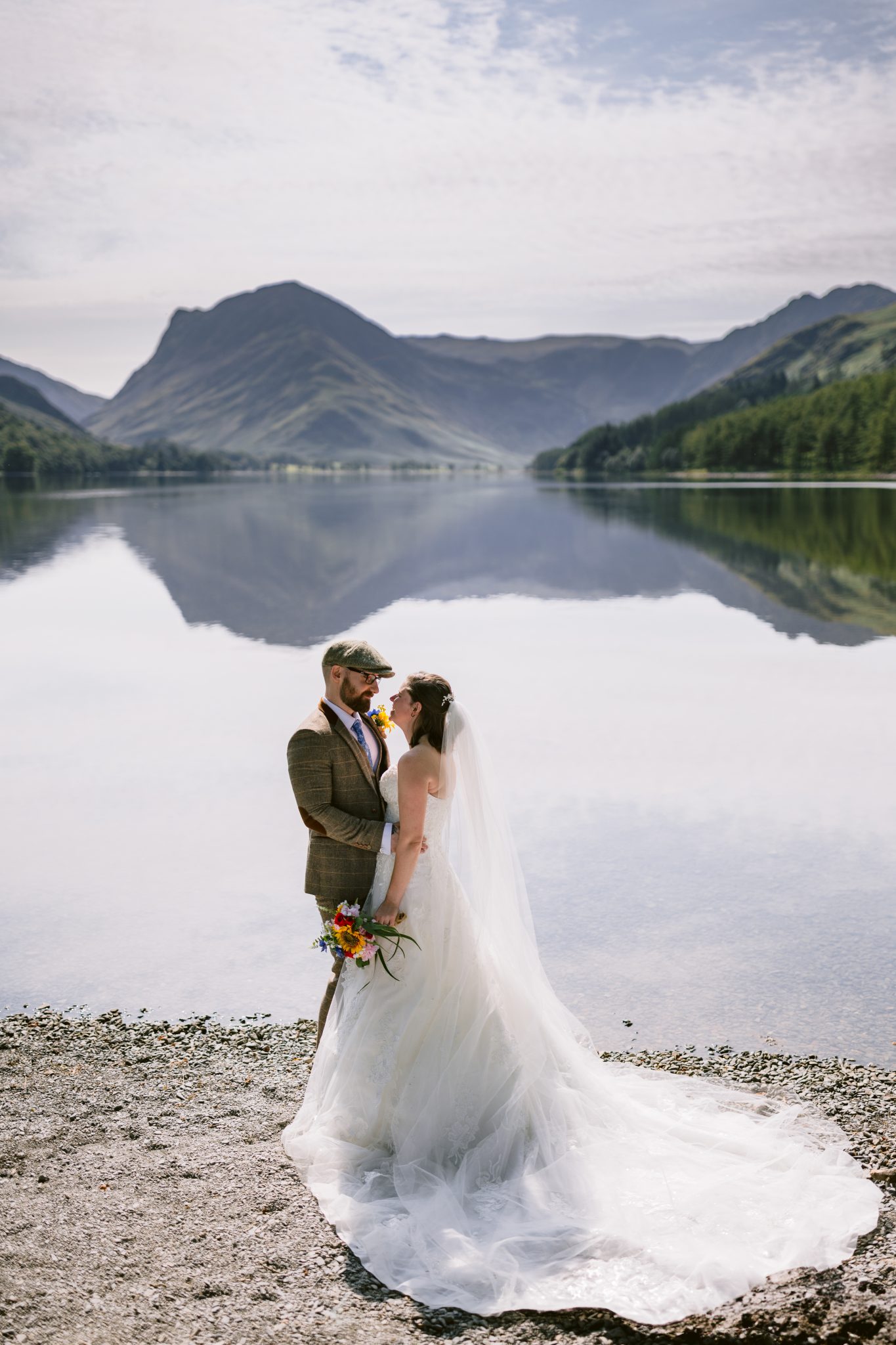 Bride and groom stand by a lakeside with mountains in the background. The bride holds a bouquet. The water reflects the landscape and sky.