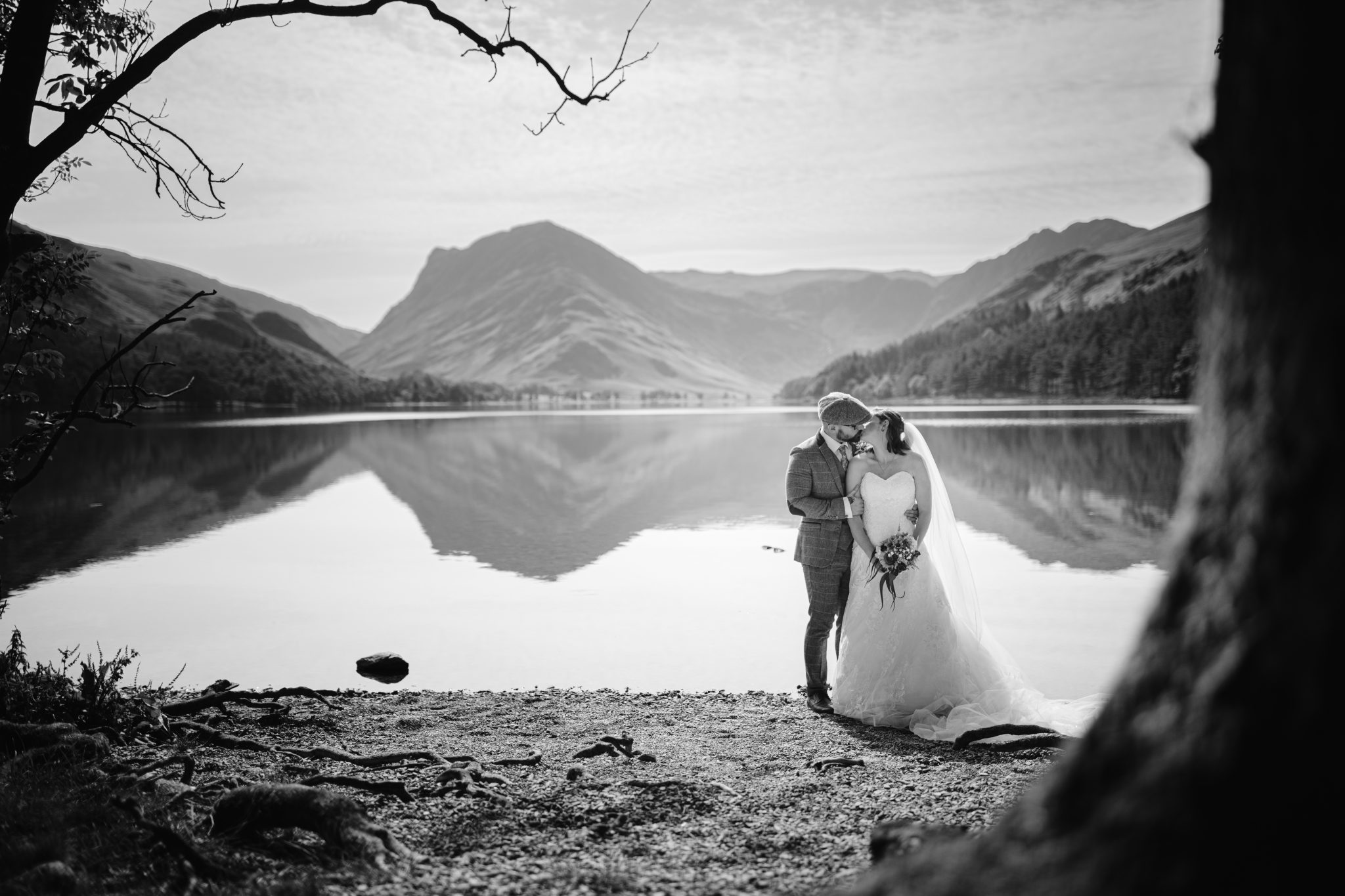 Bride and groom embrace by a lakeside, surrounded by mountains and trees, with the water reflecting the landscape.