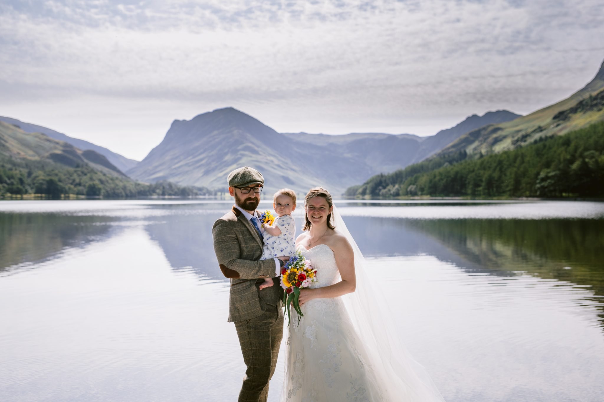 A couple in wedding attire stands by a lake, holding a child. Mountains and trees are in the background under a cloudy sky.