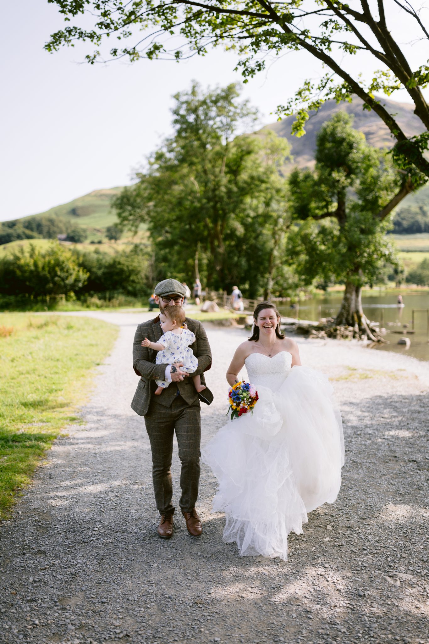 Man in a suit holding a baby and a woman in a white wedding dress walking on a path near a lake and trees.