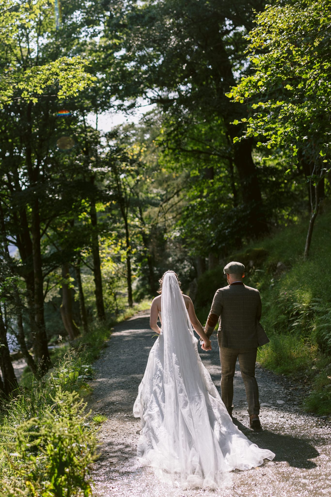 Bride and groom holding hands, walking down a tree-lined path in sunlight.