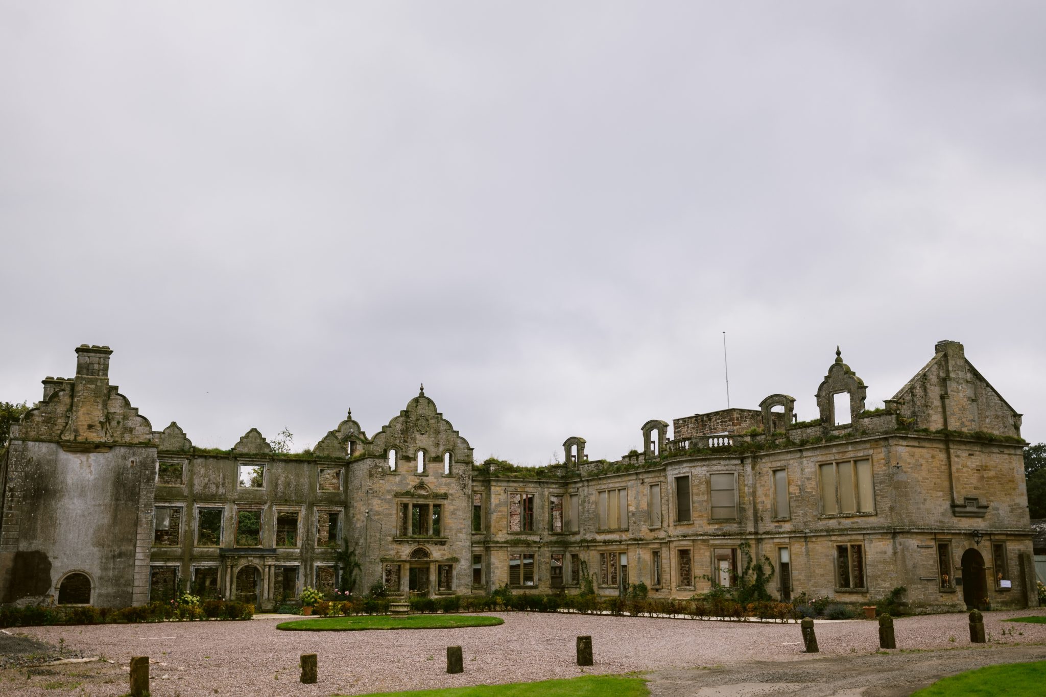 The old, weathered stone building stands in ruins under a cloudy sky, surrounded by lush gardens and a grassy expanse with a gravel path. Kirklinton Hall’s storied past adds an air of mystery to this enchanting place.
