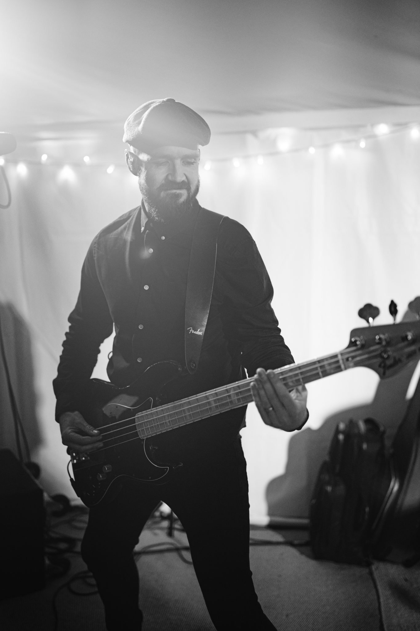 A bearded man wearing a cap plays an electric bass guitar in a dimly lit room with string lights, reminiscent of a cozy corner at Kirklinton Hall.