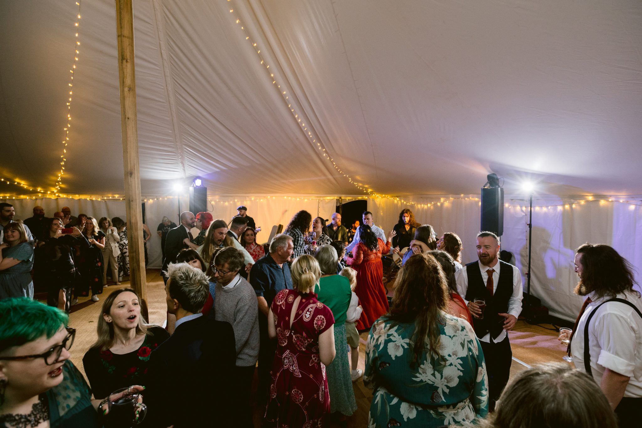 Guests dancing and socializing at a lively indoor event under the enchanting white tent ceiling of a Summer Marquee Wedding, with string lights illuminating the Gardens of Kirklinton Hall.