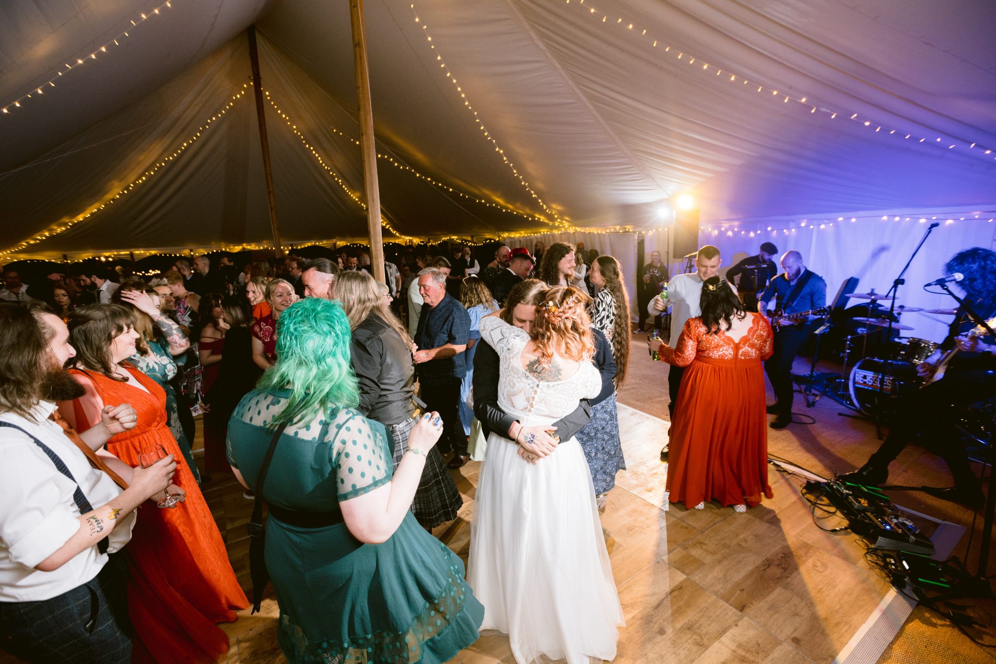 At a Summer Marquee Wedding in the enchanting gardens of Kirklinton Hall, a bride and groom dance under twinkling string lights. Surrounded by joyous guests, the couple sways to the melodies of a live band, filling the tent with a magical ambiance.
