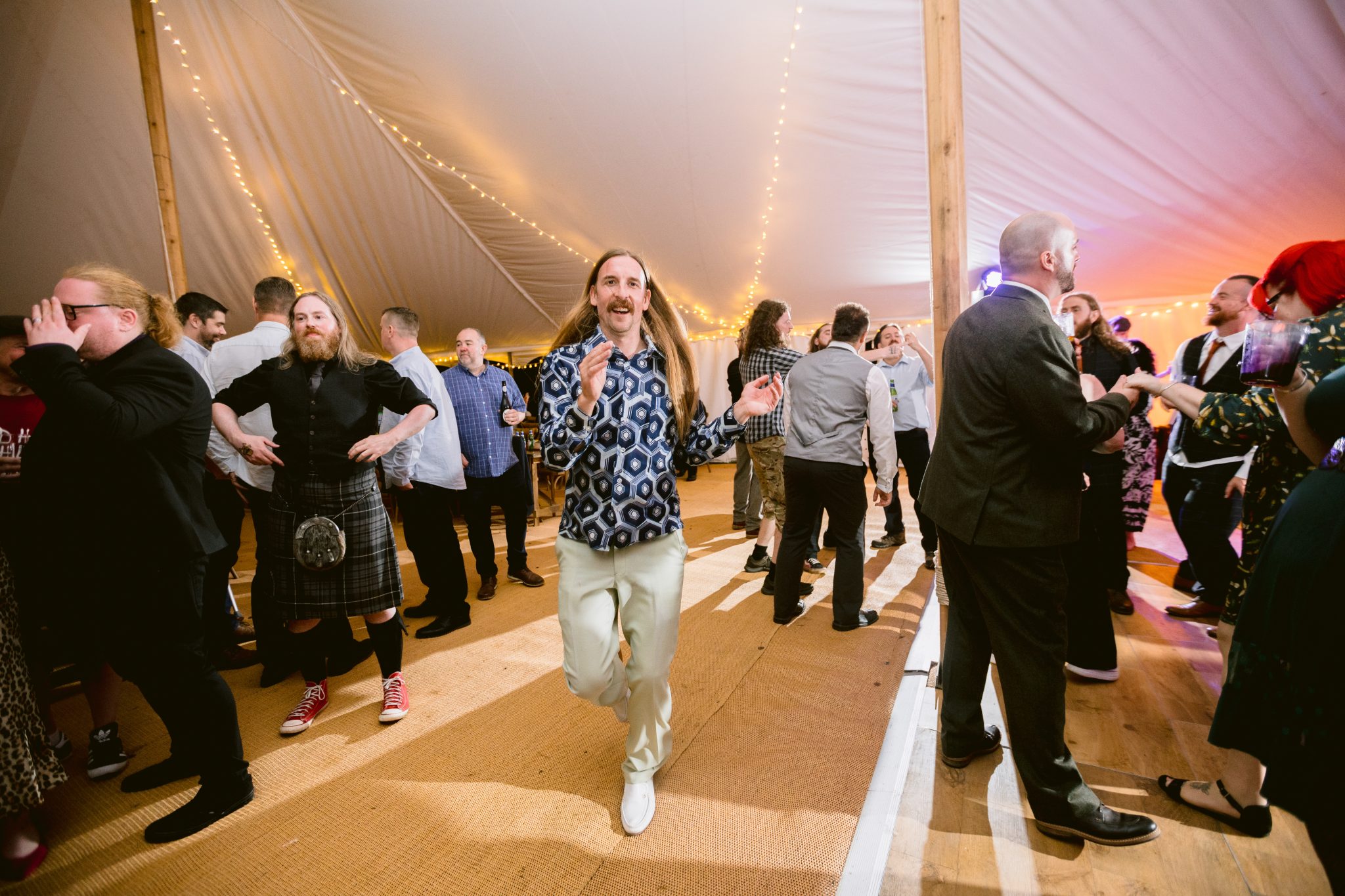 A man with long hair and a patterned shirt dances energetically in a tented venue at Kirklinton Hall, while others stand and converse in the background, celebrating the lively wedding.