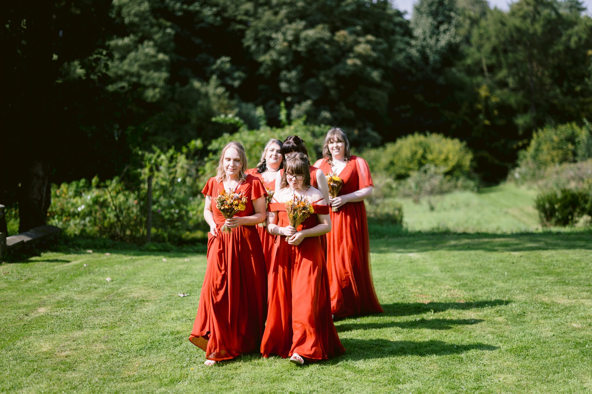 Four women in red dresses walk across a grassy field at Kirklinton Hall, each holding a bouquet of flowers. The scene, perfect for a summer marquee wedding, is framed by the lush trees and greenery visible in the background.