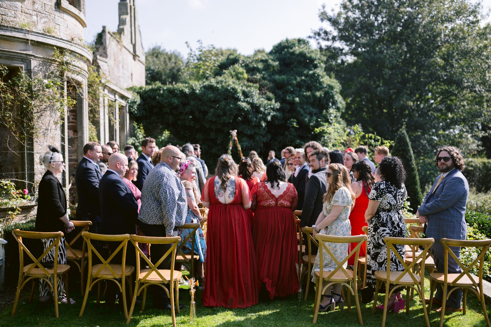 Guests gathered for a beautiful outdoor ceremony, standing and watching the couple exchange vows amid the lush greenery of Kirklinton Hall. Rows of wooden chairs lined the gardens, offering a stunning backdrop with a stone building's timeless charm.