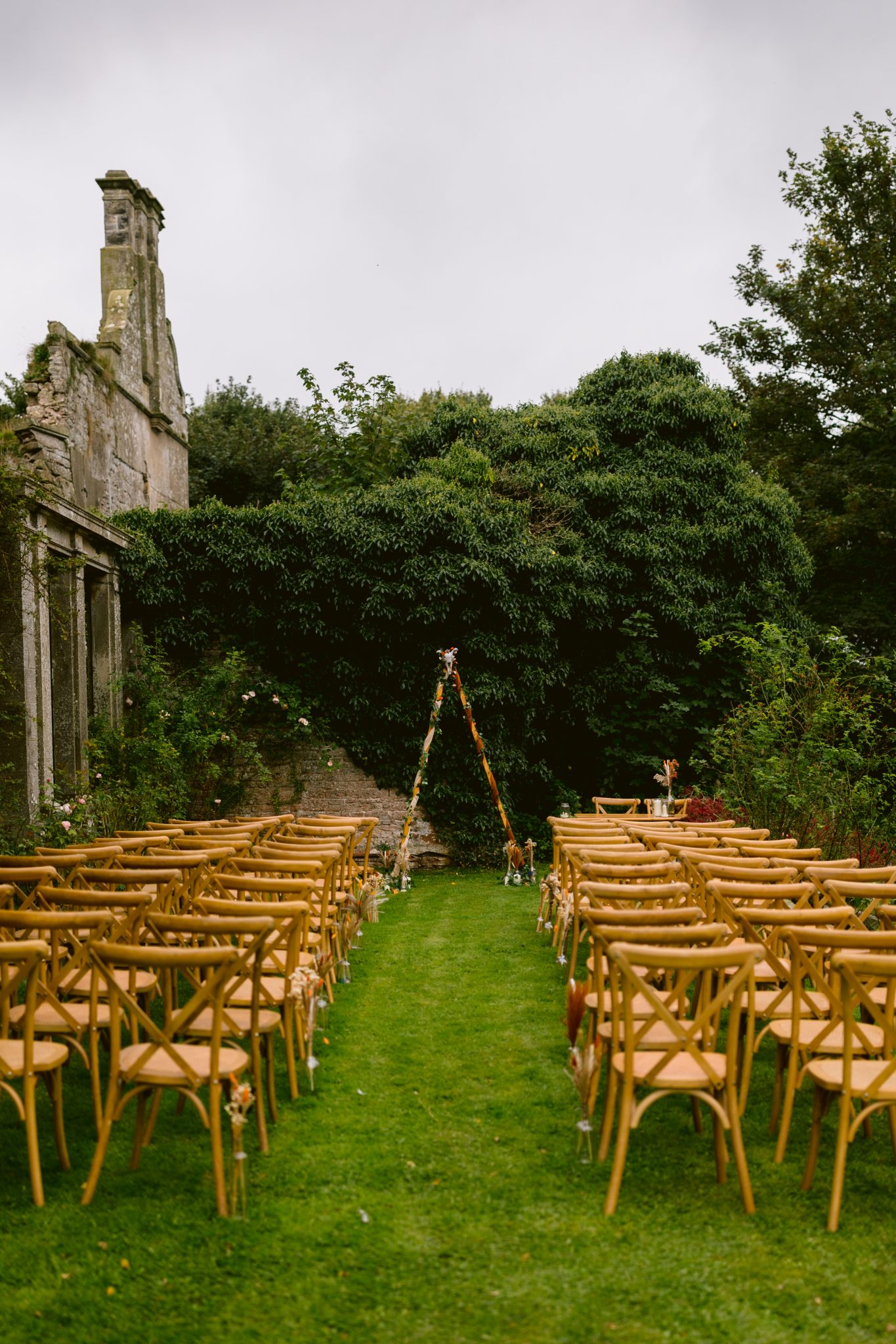 A charming outdoor wedding setup at Kirklinton Hall features rows of wooden chairs facing a small floral arch, nestled among lush gardens and an old stone structure in the background, perfect for a summer marquee wedding.
