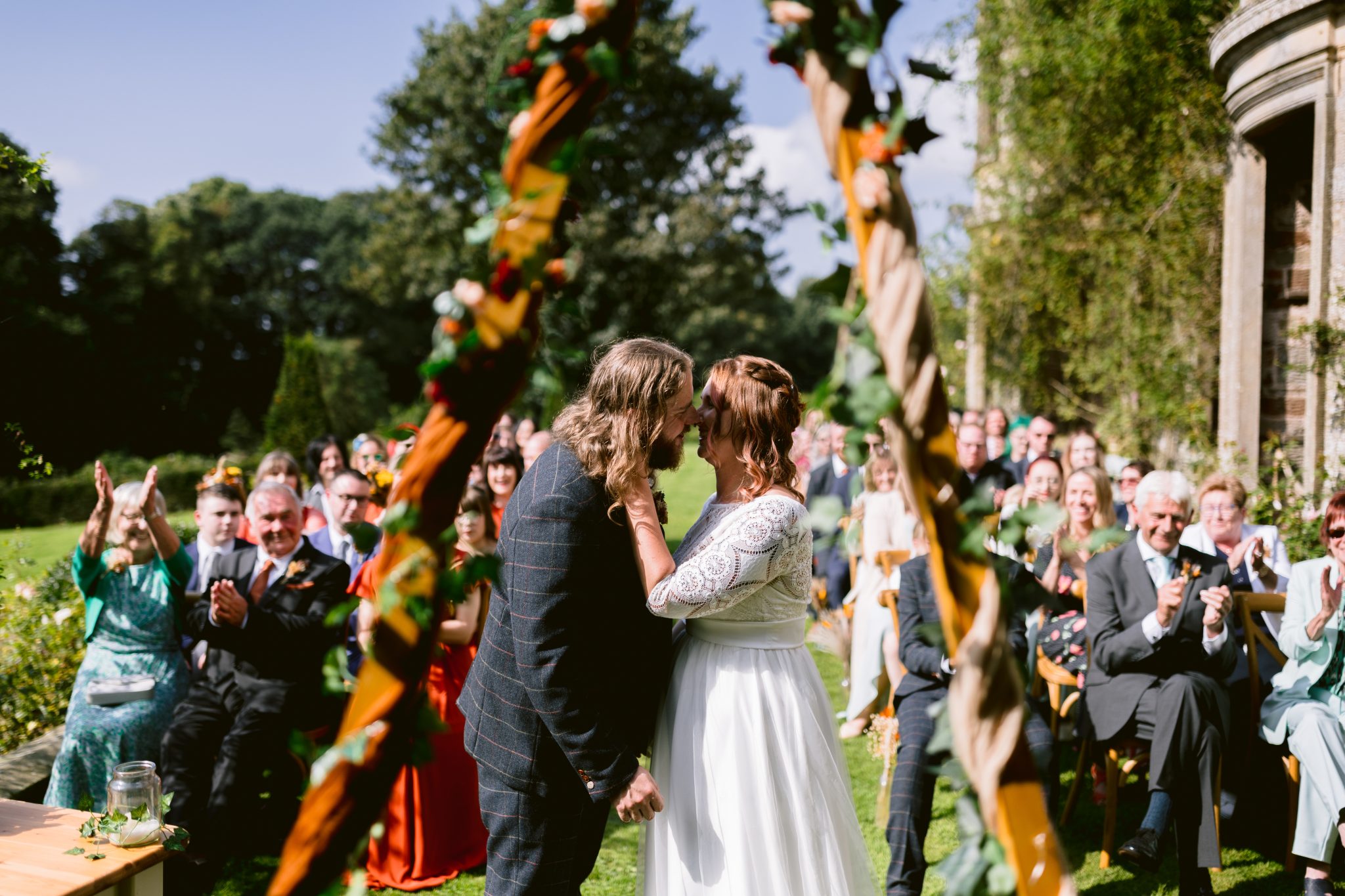 Amidst the enchanting gardens of Kirklinton Hall, a bride and groom share a kiss during their summer marquee wedding. Guests, seated on benches adorned with decorative ribbons, witness this beautiful outdoor ceremony.