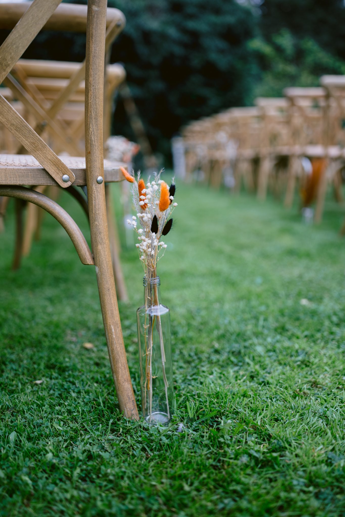A glass bottle with dried flowers sits beside a wooden chair on the lush green grass, evoking the charm of Kirklinton Hall. Rows of wooden chairs in the background suggest a picturesque setting perfect for a summer wedding.
