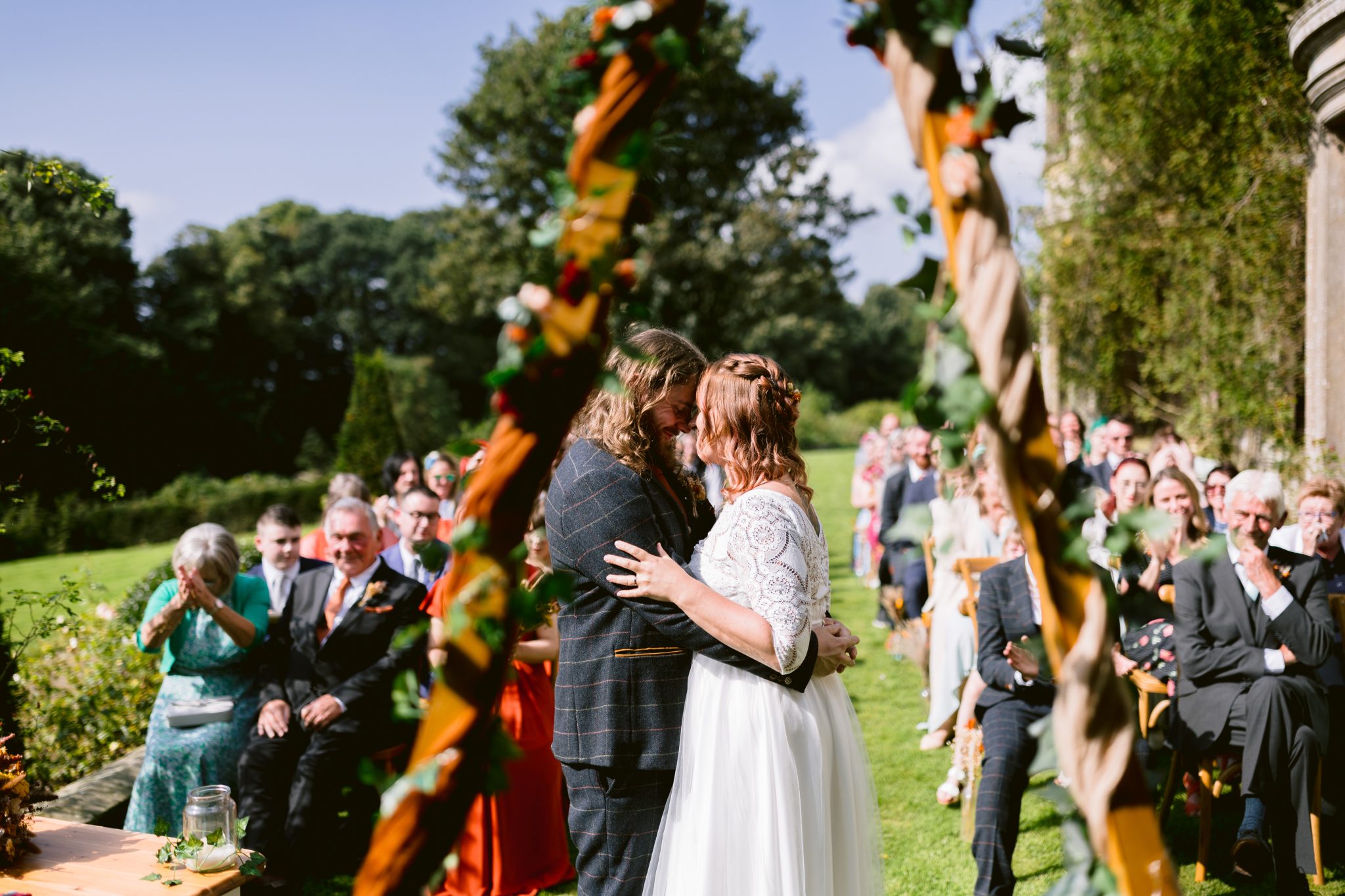 Two people in wedding attire stand close amidst guests on a sunny day at Kirklinton Hall. Surrounded by lush trees and vibrant greenery, it's the perfect setting for a summer marquee wedding in the beautiful gardens.