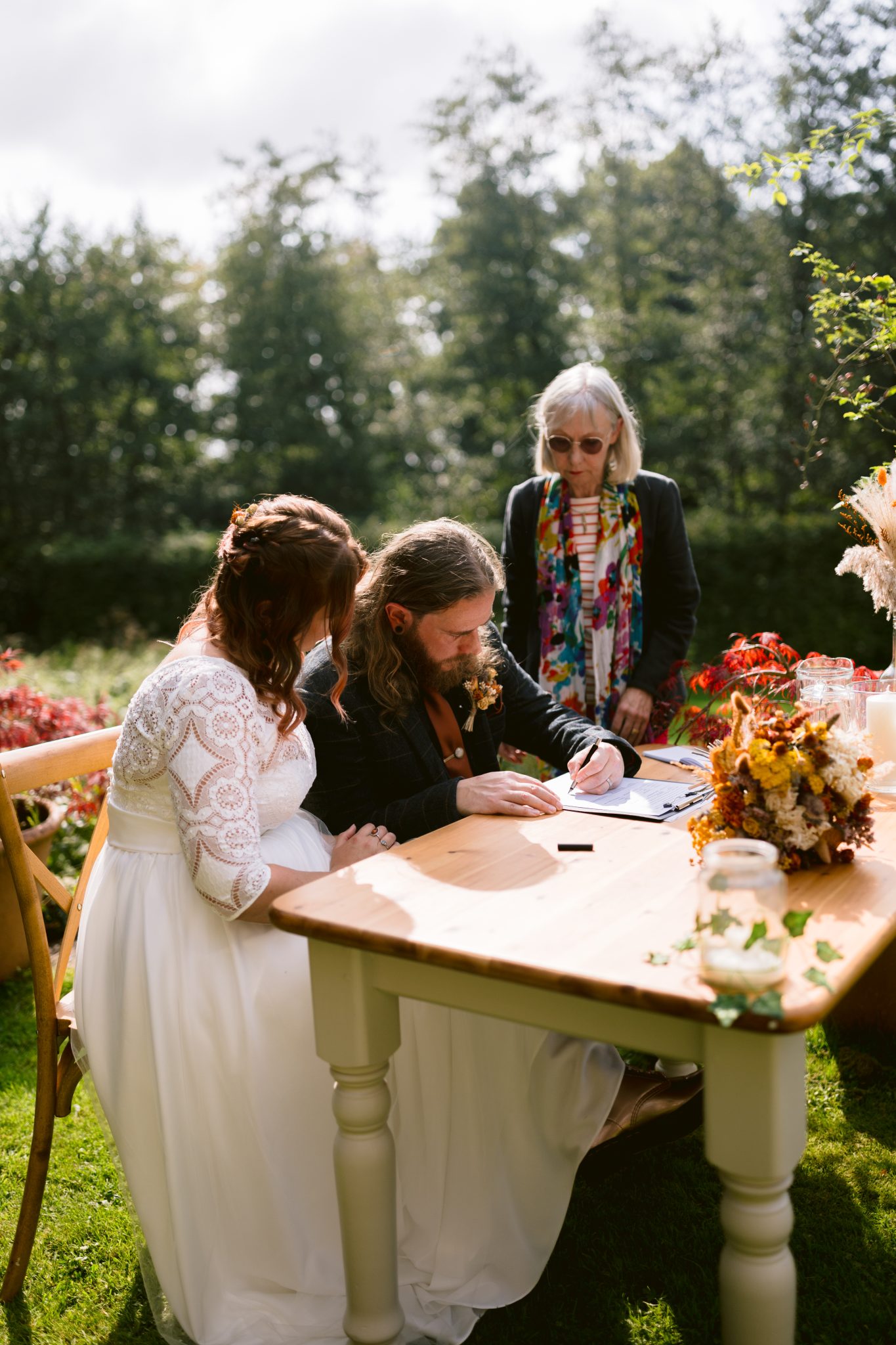 At a summer marquee wedding in the enchanting gardens of Kirklinton Hall, a bride and groom sign documents at an outdoor table adorned with flowers, while an officiant stands nearby.